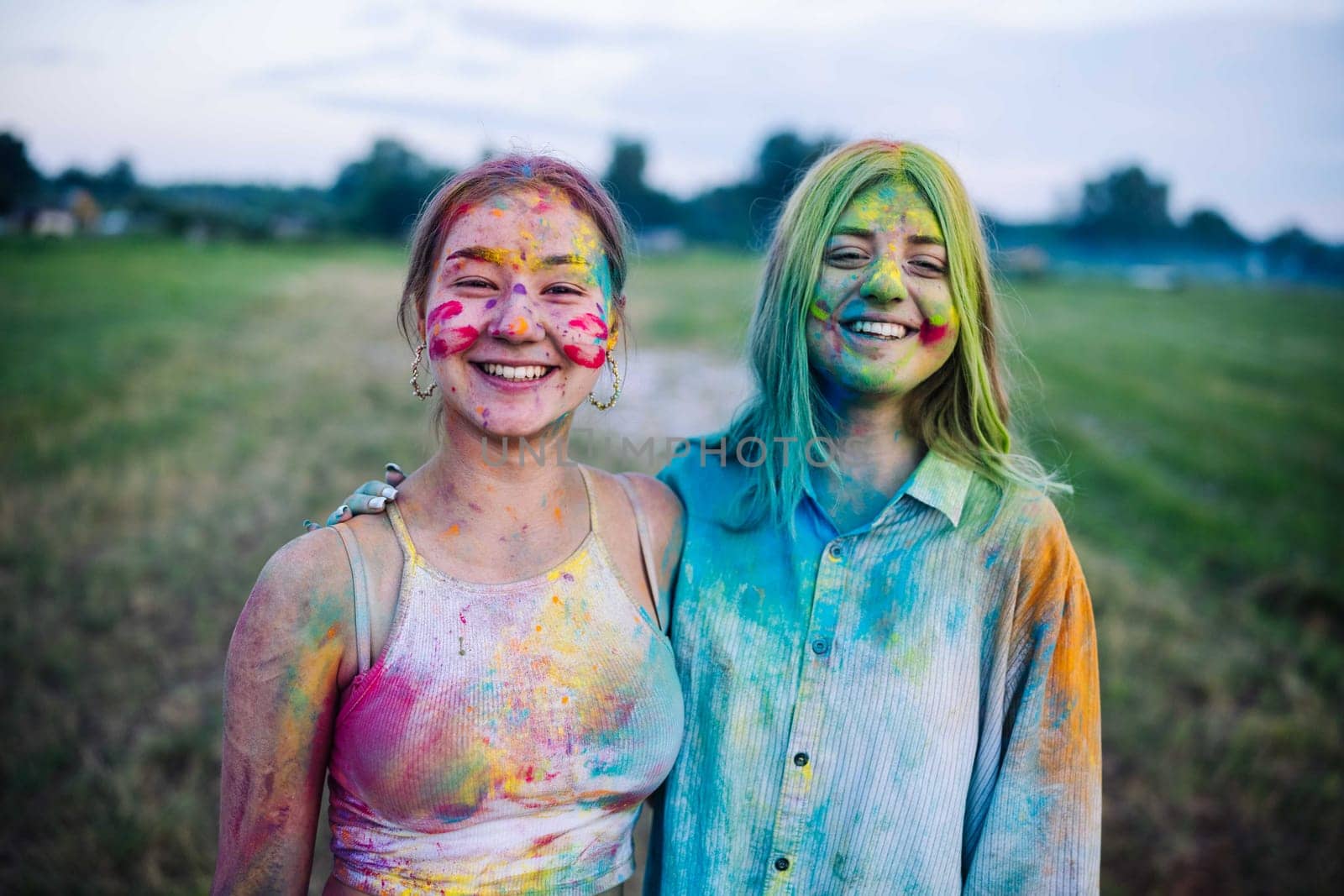Two vibrant young women, coated in colorful powder, embrace and share smiles at the holi festival festivities