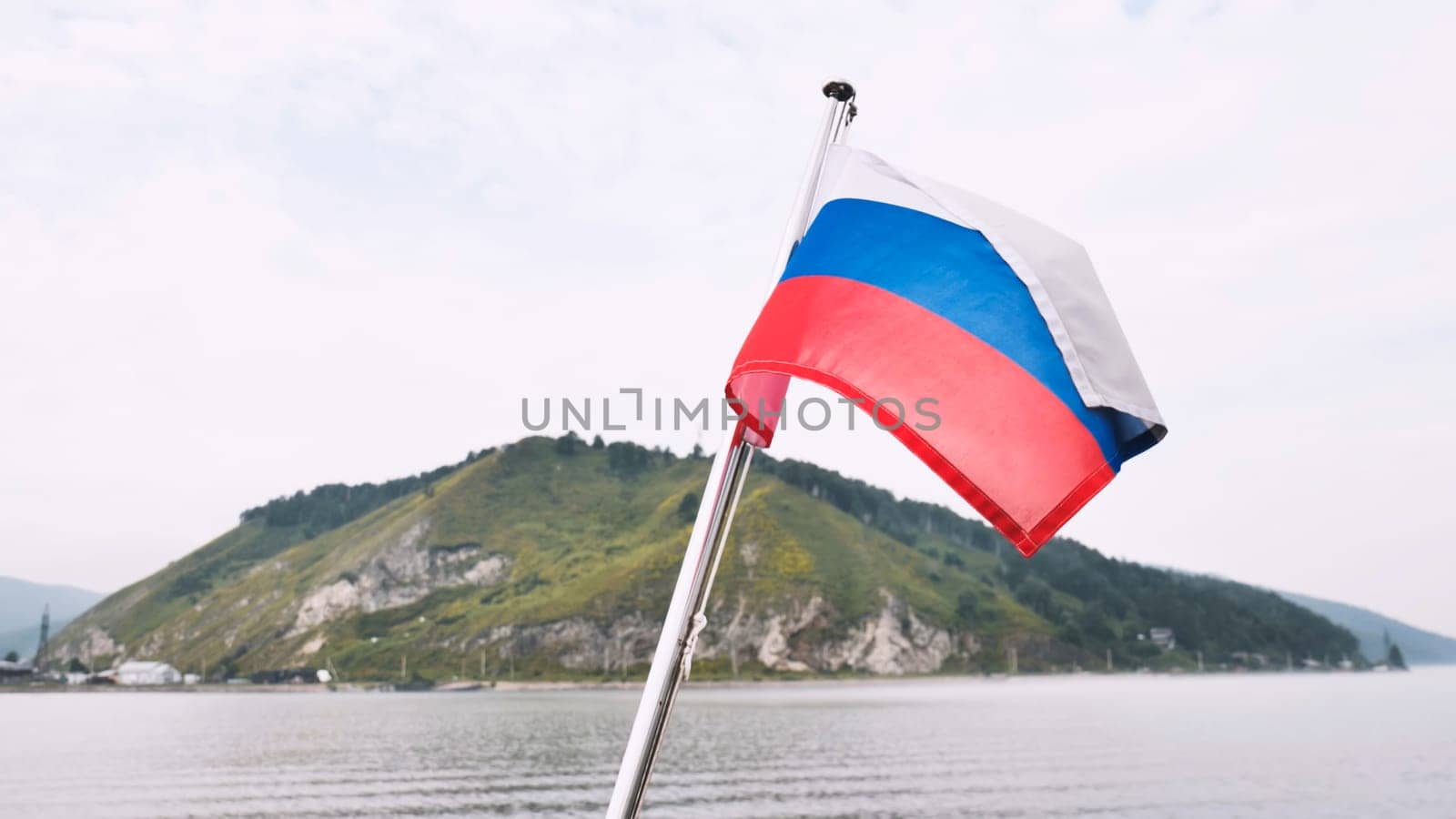 Russian flag waving on boat sailing volga river, green riverbank rising, cloudy atmospheric sky stretching across horizon