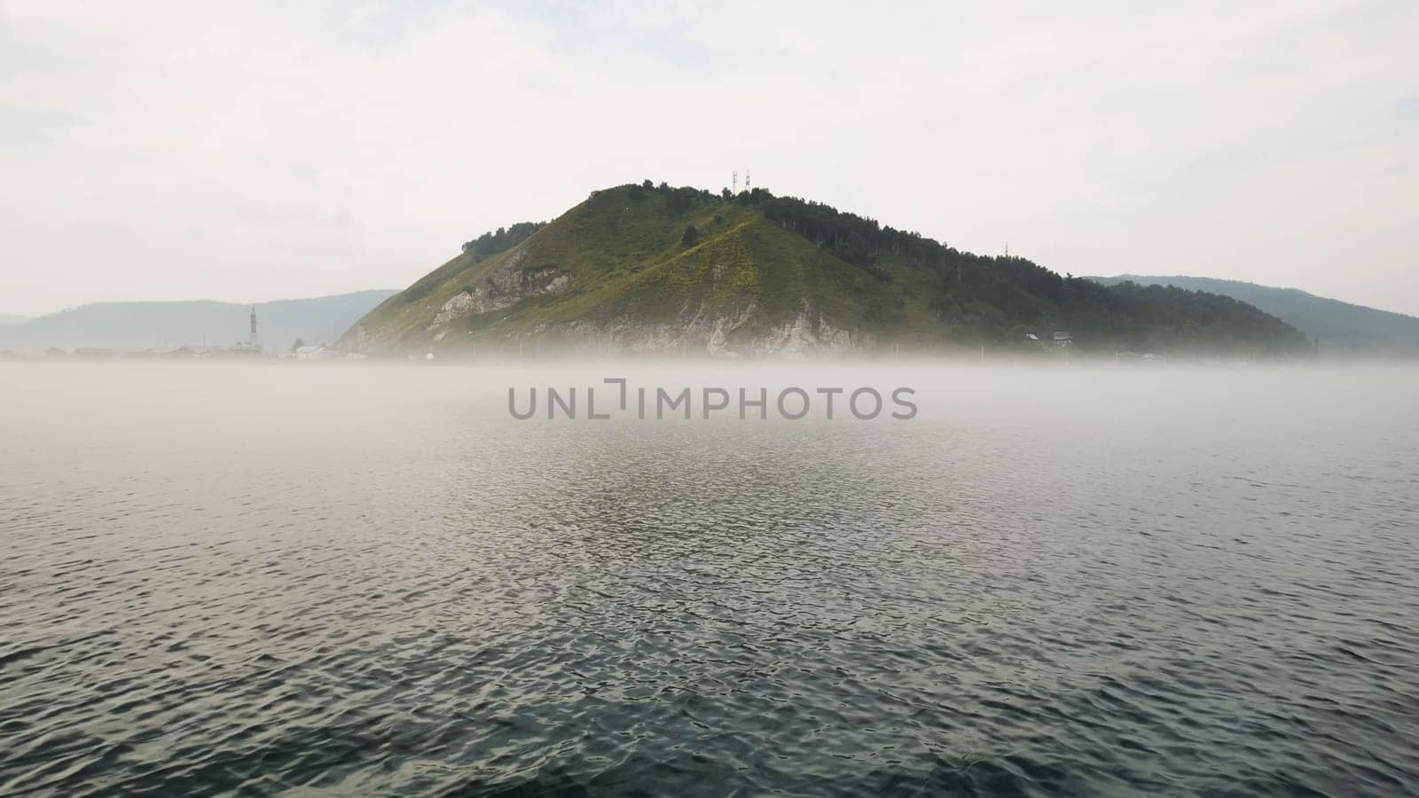 Scenic view of lake baikal with fog partially obscuring a small island featuring telecommunication towers, showcasing the serene beauty and mystery of nature