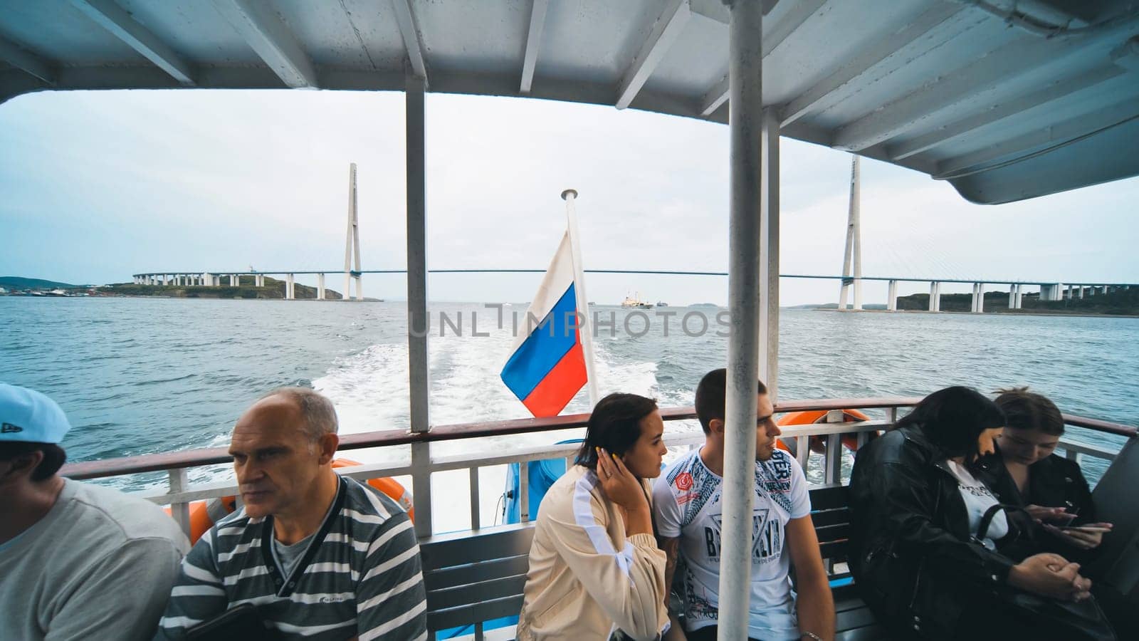 Tourists enjoying a ferry trip under the russian bridge in vladivostok, russia, with the russian flag waving in the foreground