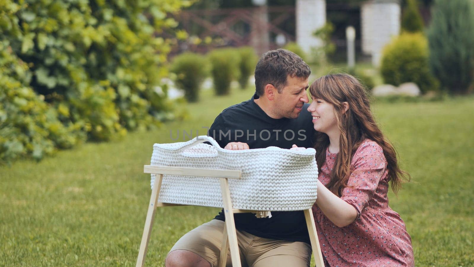 Loving parents gazing adoringly at sleeping newborn nestled peacefully in wooden crib under soft natural sunlight near garden setting