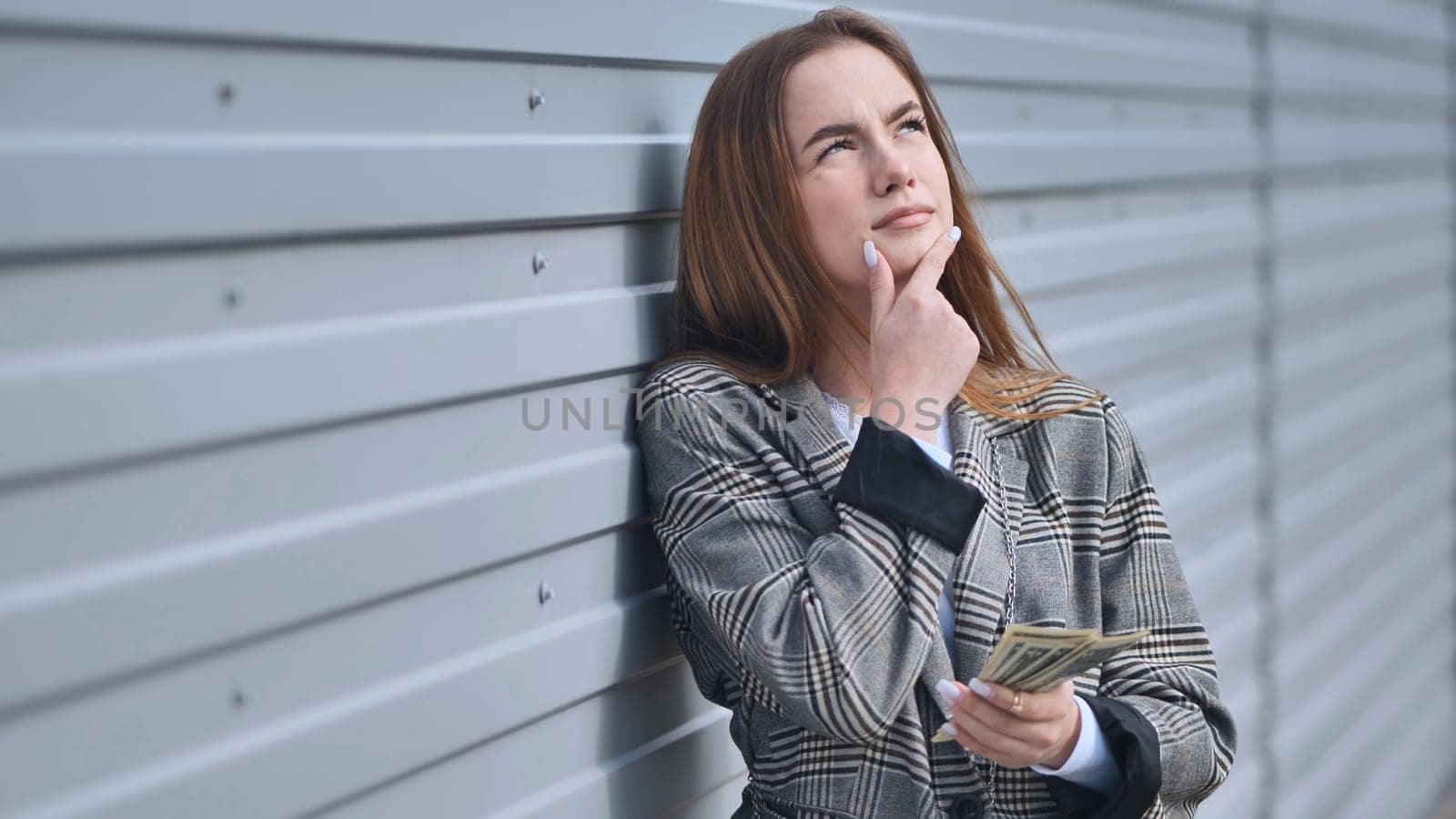 Pensive businesswoman holding us dollar banknotes, leaning against a corrugated iron wall and thinking about investments