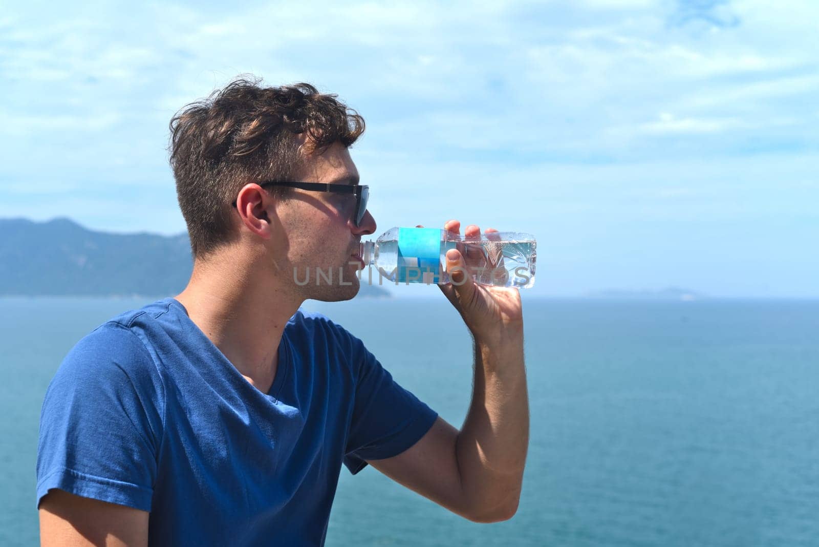 Young man drinking water from a bottle against sea and blue sky