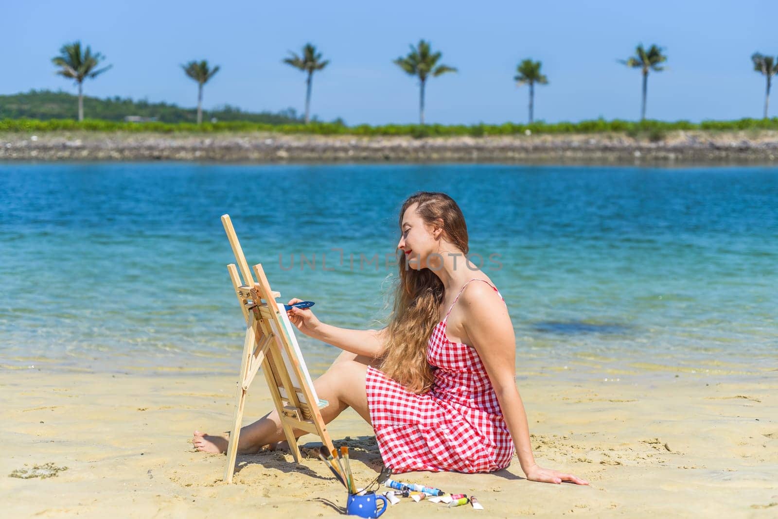 Young artist woman sitting at the beach with a jar with paint brushes and easel close to sea and palms in summer