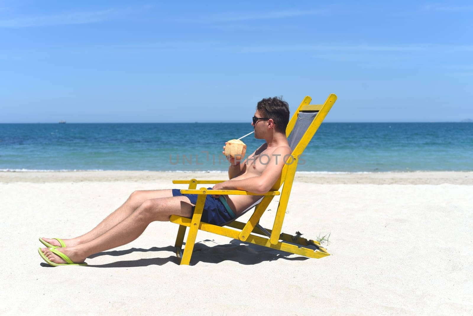 Young happy man drinks coconut juice sitting on deck chair on a south china sea beach in vietnam