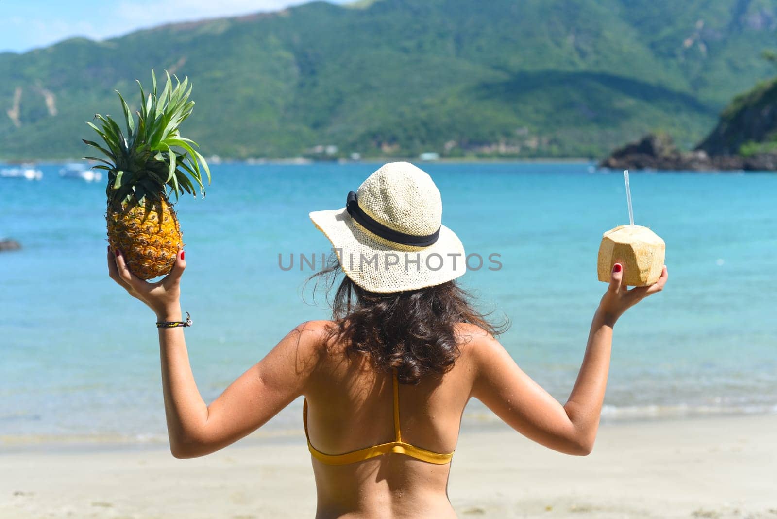 Young woman in yellow swimsuit holding coconut and pineapple on sandy beach