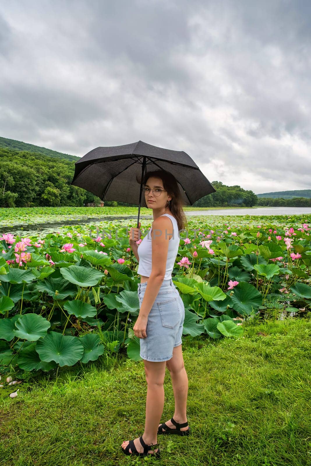 Upset woman holding blak umbrella under rain and enjoying rainy weather near lotus lake in the Far East of Russia