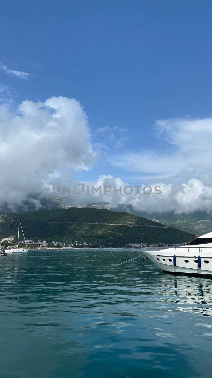 A tranquil harbor with yachts docked, clear reflections on the water, and mountains under a bright blue sky with fluffy clouds.