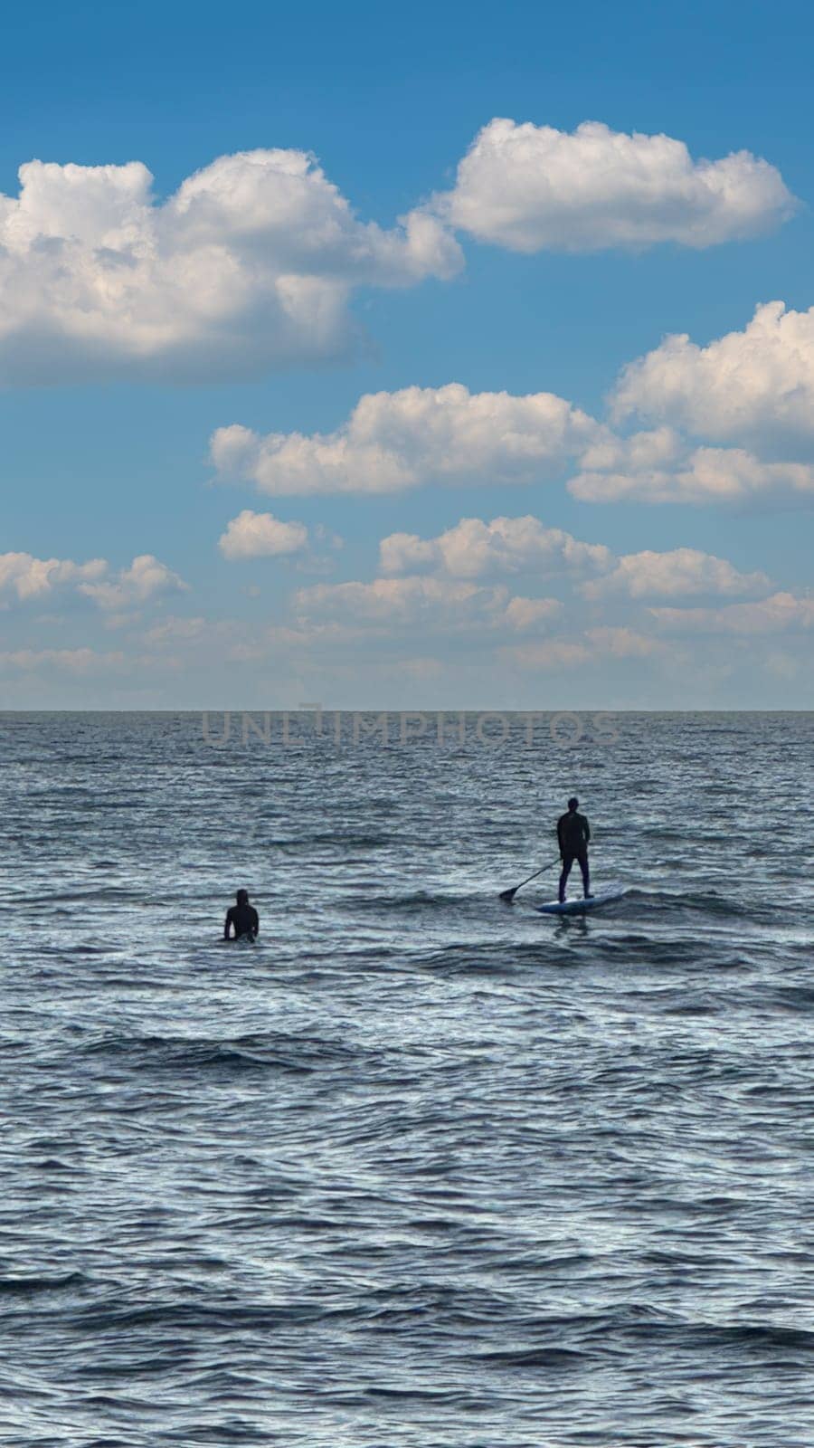Silhouette of two people on a stand up paddle board in calm open water under a bright blue sky with fluffy clouds.