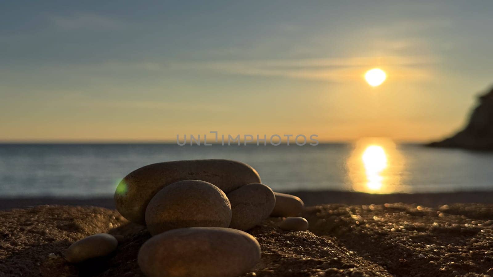 Smooth rocks on a beach with the sun setting over calm waters, a warm glow reflecting on the sea. High quality photo
