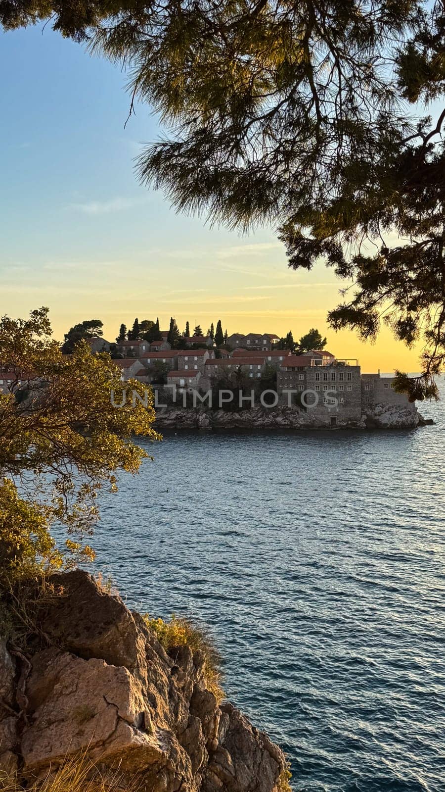 Sveti Stefan, a small island and resort in Montenegro.Stone buildings and lush greenery, view from a rocky shore framed by pine trees at sunset.