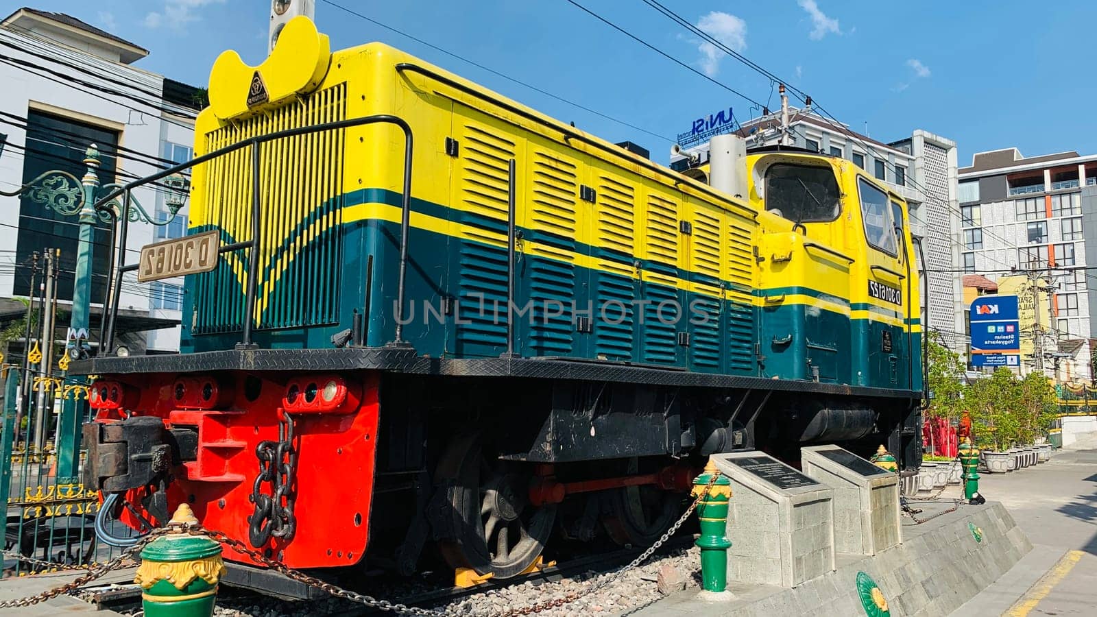 Bright yellow and green antique Diesel locomotive D30162 on display used to be maintained by Kereta Api Indonesia, showcasing railway history in a city setting. Features urban surroundings, clear blue skies, and represents both transportation innovation and nostalgia for vintage trains.