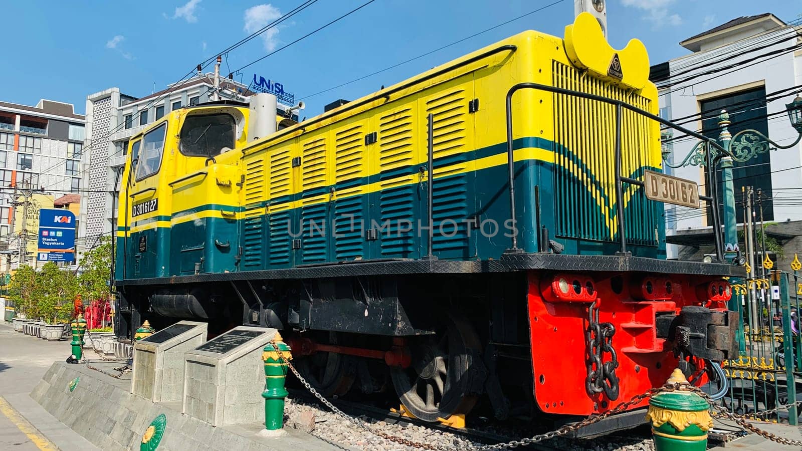 Bright yellow and green antique Diesel locomotive D30162 on display used to be maintained by Kereta Api Indonesia, showcasing railway history in a city setting. Features urban surroundings, clear blue skies, and represents both transportation innovation and nostalgia for vintage trains.