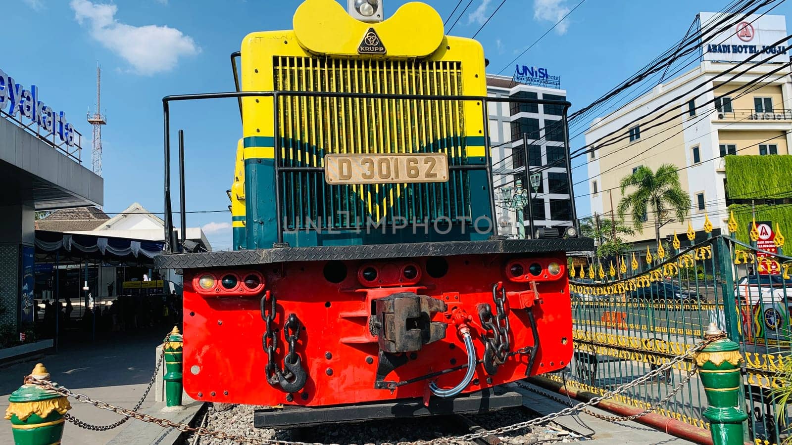 Bright yellow and green antique Diesel locomotive D30162 on display used to be maintained by Kereta Api Indonesia, showcasing railway history in a city setting. Features urban surroundings, clear blue skies, and represents both transportation innovation and nostalgia for vintage trains.