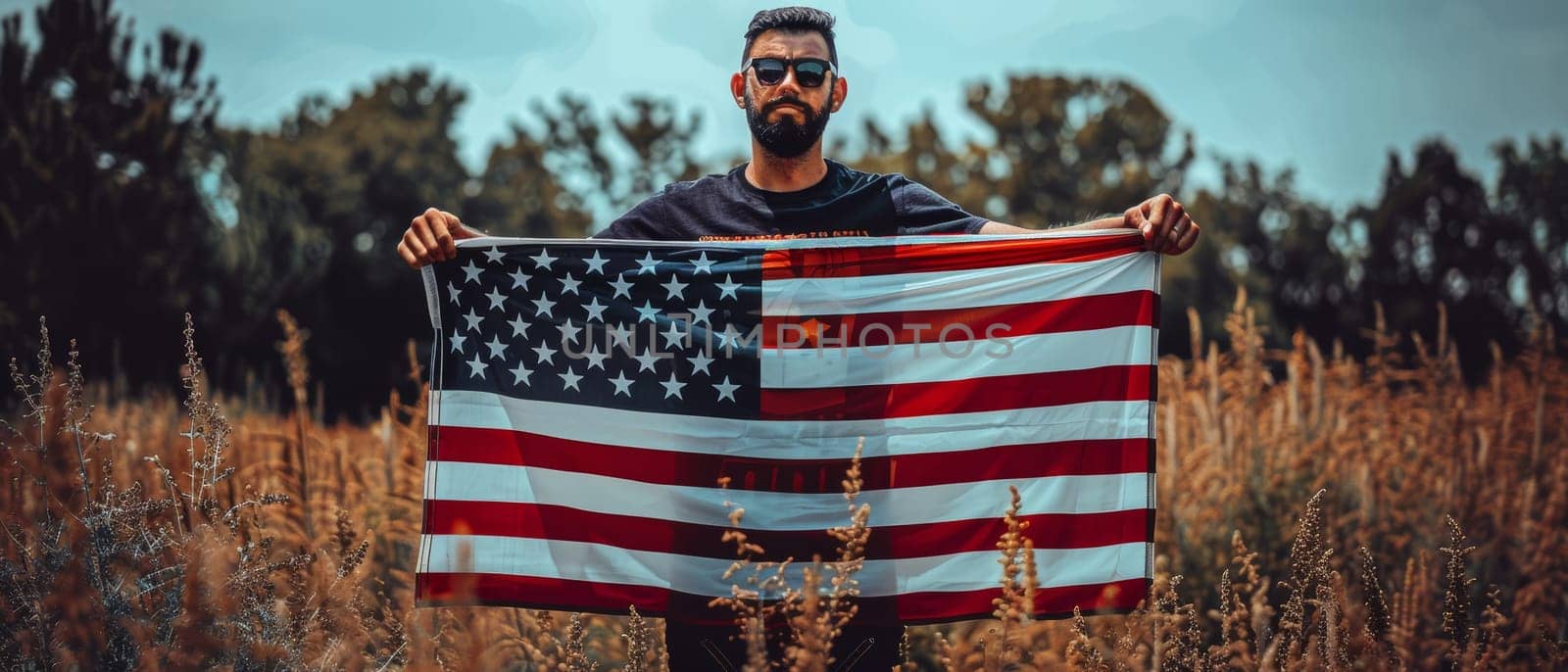 A bearded man in sunglasses and a dark t-shirt proudly holds the American flag, with a background of golden wild grass under a clear sky