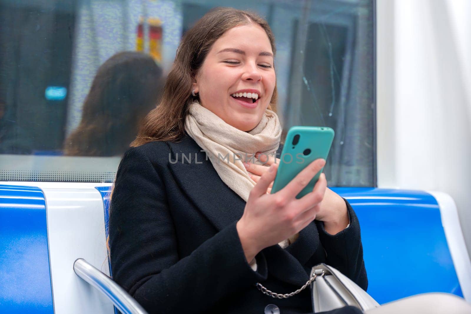 Cheerful young woman in a black coat and beige scarf sitting inside a modern subway train, laughing while looking at her smartphone. She enjoys a fun moment during her commute.
