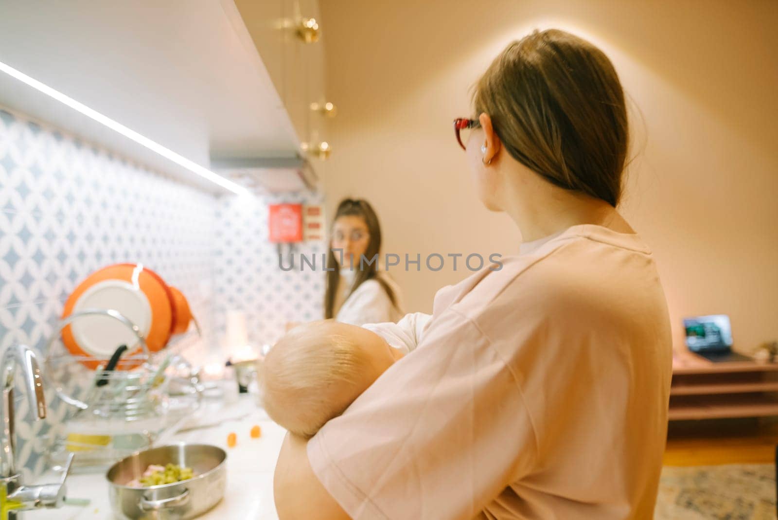 A loving mother cradles her baby while engaging in a friendly conversation with a friend in a stylish kitchen