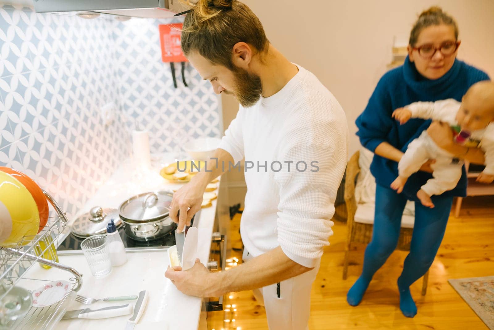 A cozy and inviting kitchen scene depicts a parent happily cooking while the other lovingly cares for a baby
