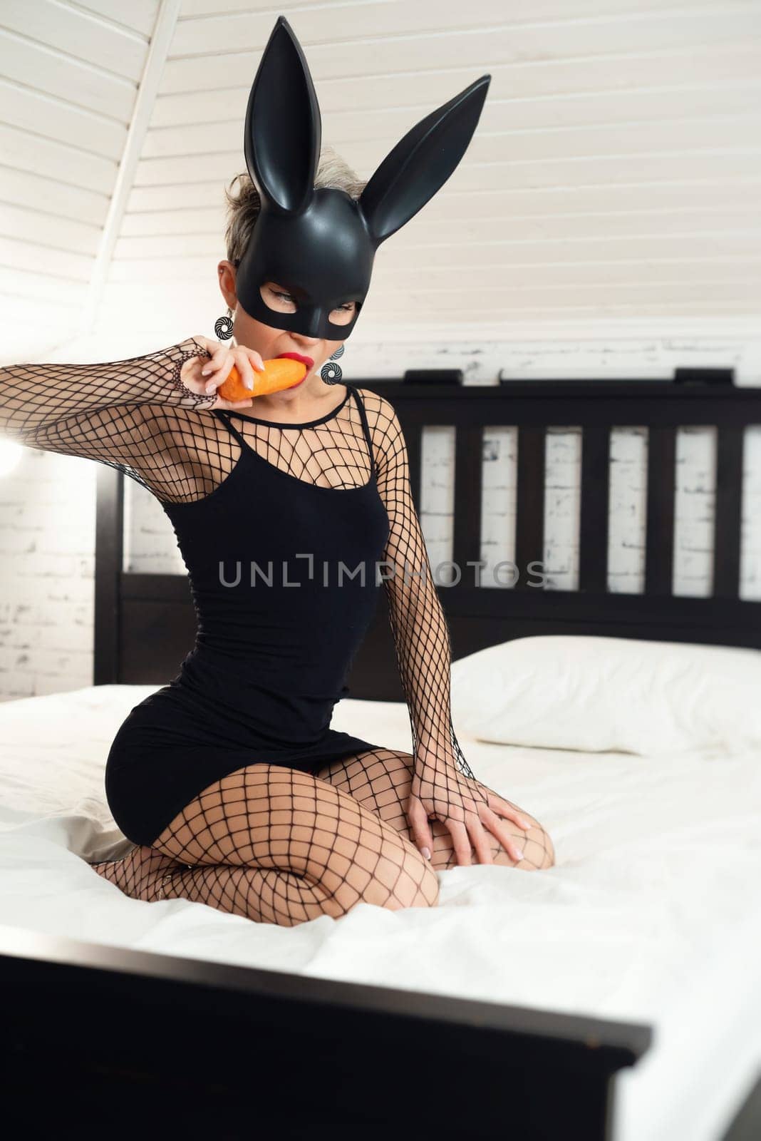 A sexy girl in a short black dress and a rabbit mask from sex shop erotically plays with carrots on her hand and in her mouth, nibbling