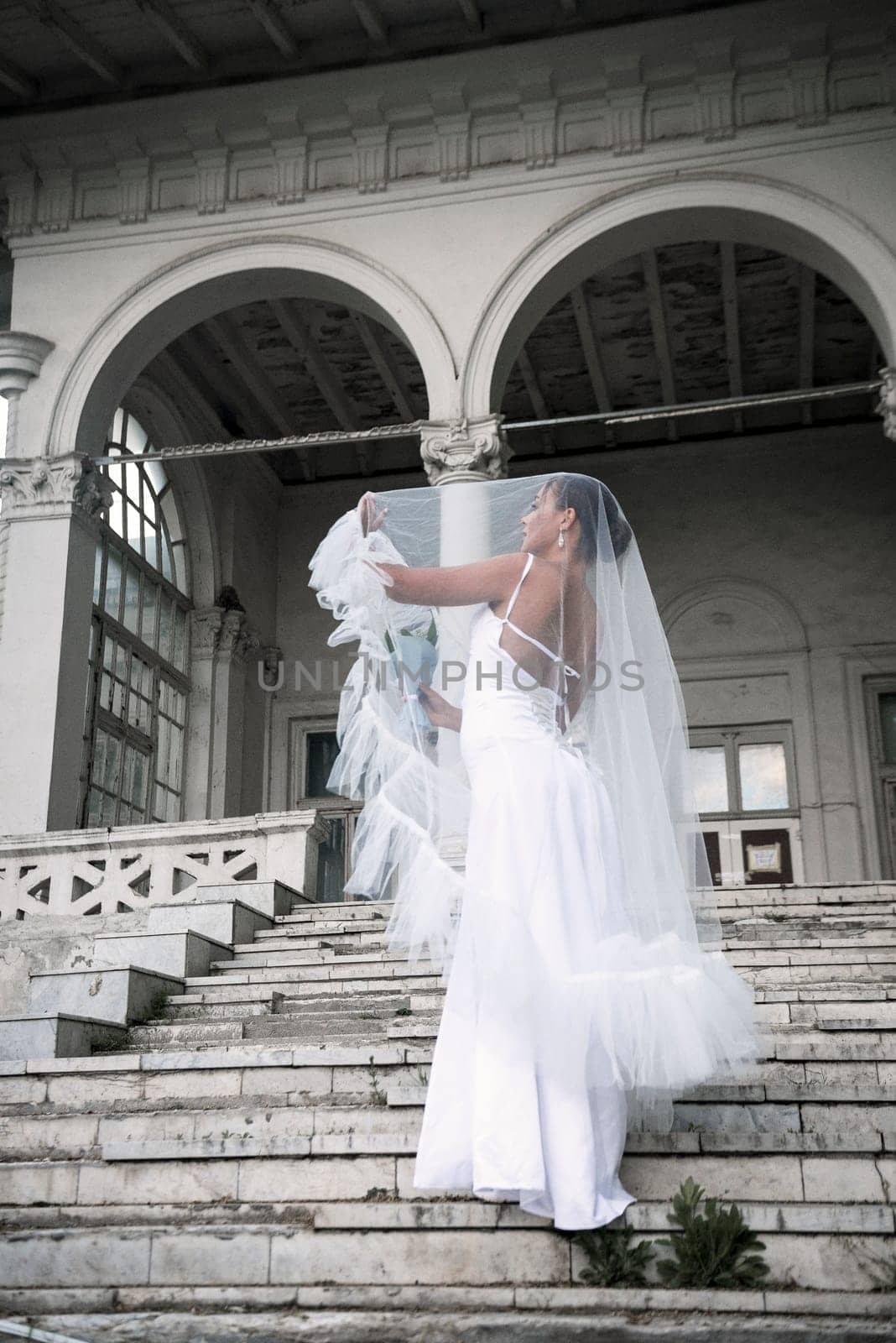 beautiful bride with a bouquet of flowers covered with a veil. a beautiful girl stands on the steps of a light building. she stands sideways in a white long dress and lifts the veil with her hands