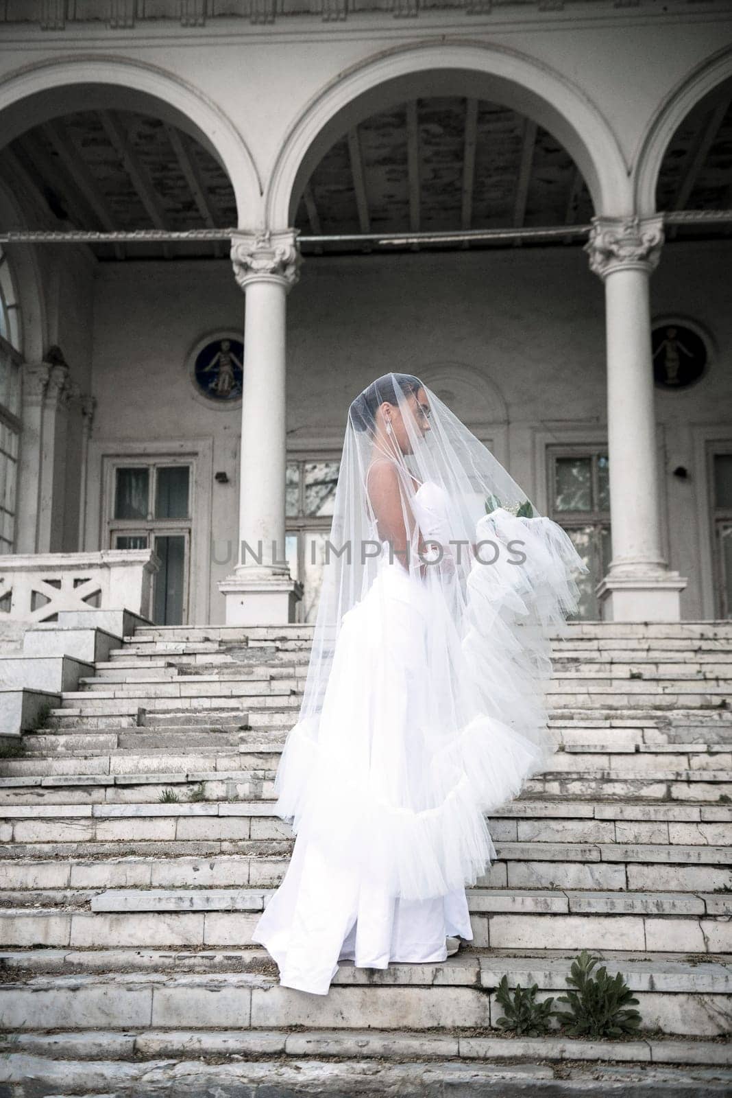 beautiful bride with a bouquet of flowers covered with a veil. a beautiful girl stands on the steps of a light building. she stands sideways in a white long dress.full length general plan