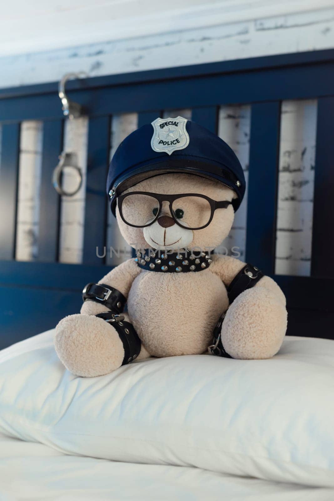 A toy teddy bear dressed in leather straps is a BDSM accessory for adult games in a police hat on the bed