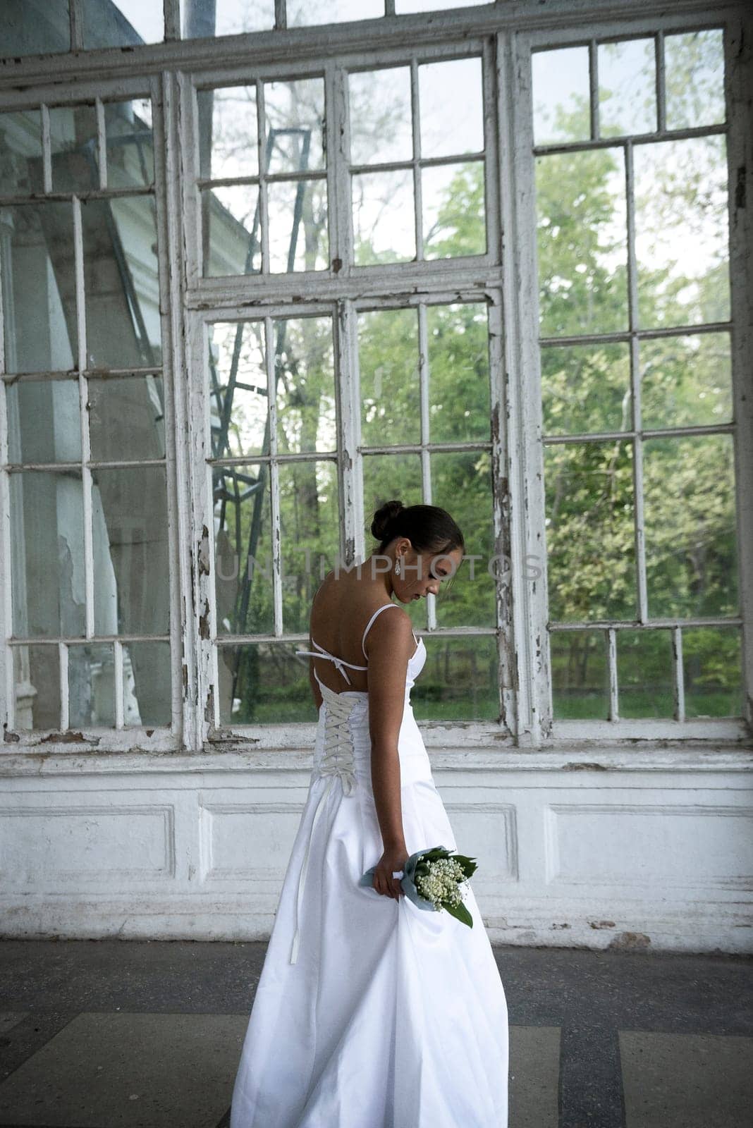 A beautiful bride in a long elegant dress with a bouquet of lilies of the valley stands near the window. A brunette girl with a beautiful hairdo, she turned around. She has white flowers