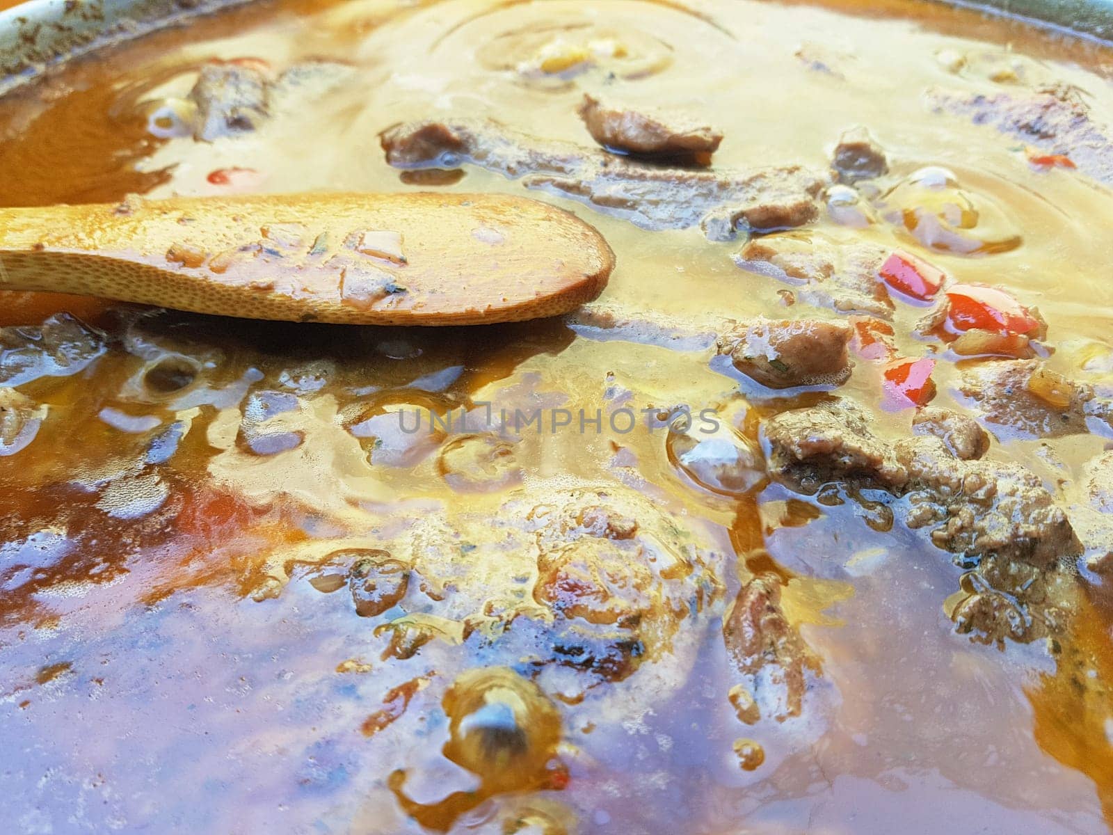 Close-up of bubbling beef stew with meat, vegetables, and rich broth in a pot. A wooden spoon rests in the liquid, showing the simmering cooking process. Rustic outdoor setting highlights traditional comfort food.