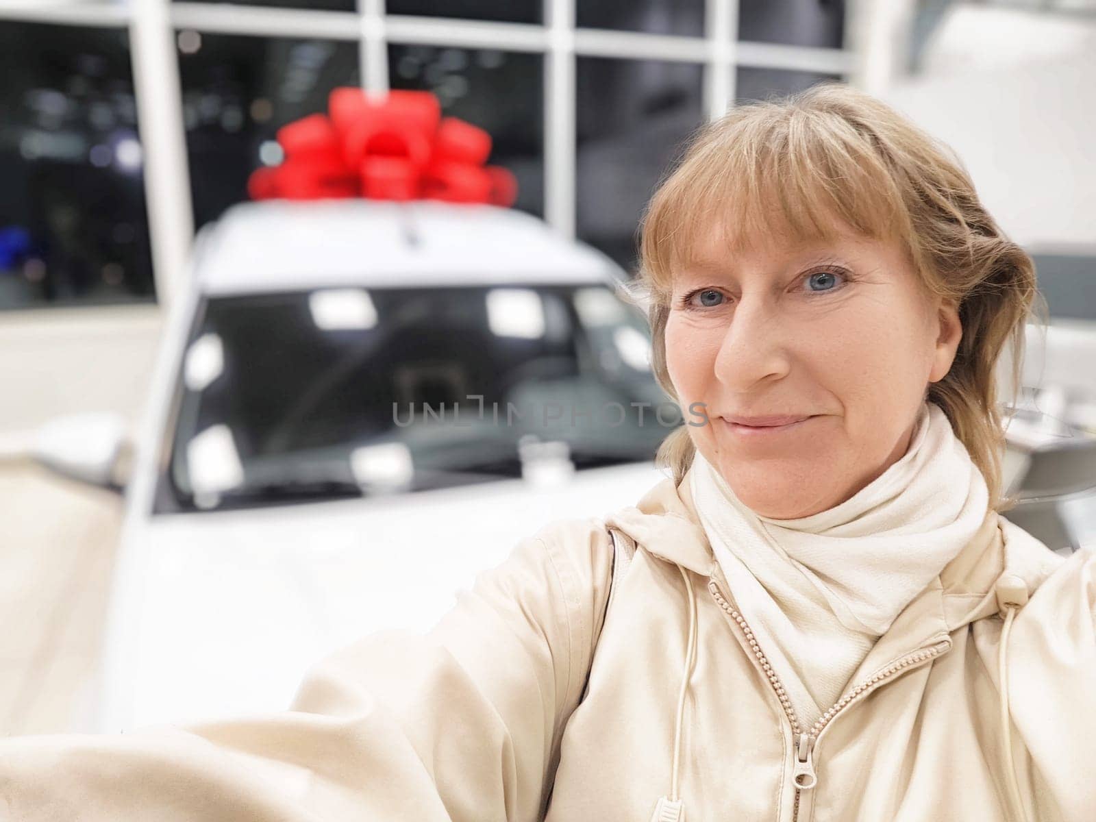 Woman takes a selfie while choosing a car with a large red bow at the dealership, excited about her purchase.