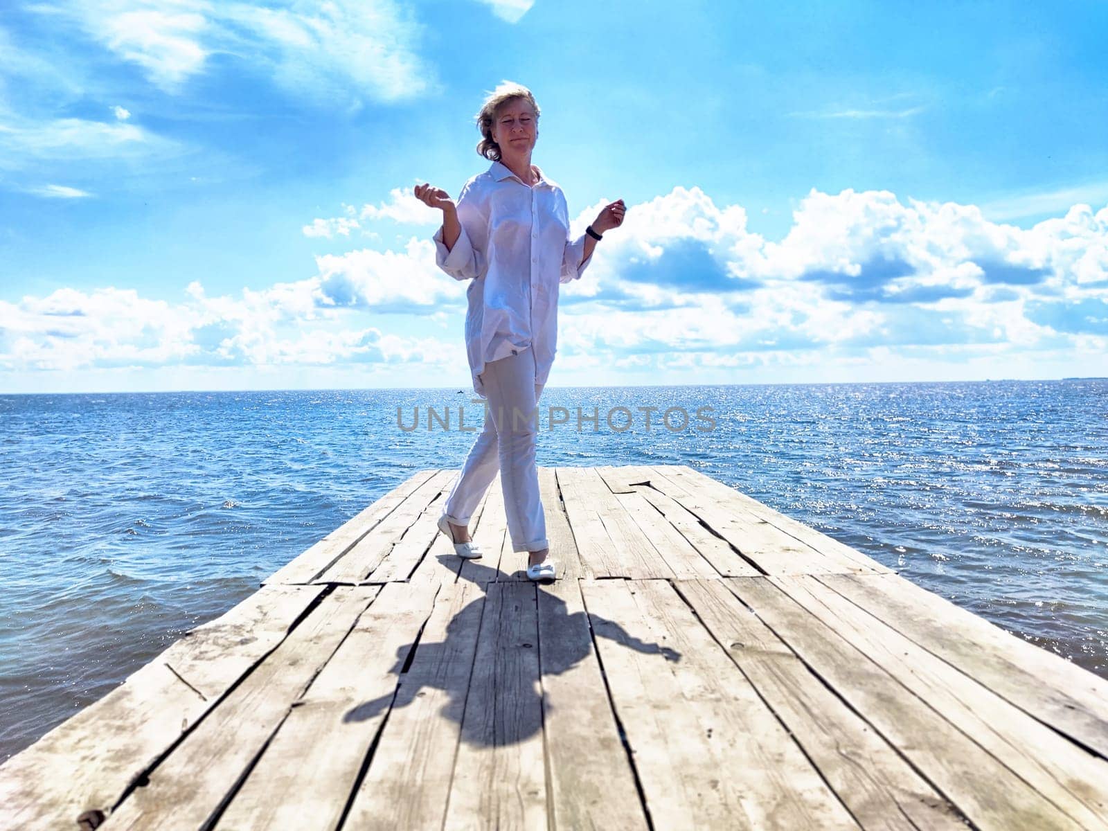 Mature adult female blogger walks on a wooden dock by the water, embracing nature on her journey under a bright sky.