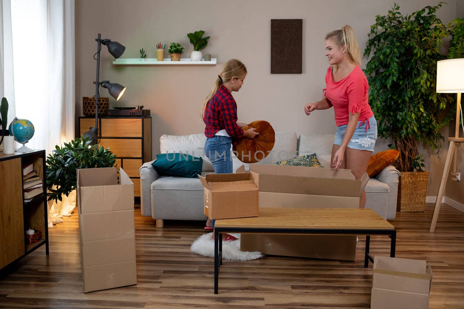 A mother and her child are happily unpacking boxes in a cozy living room filled with vibrant plants