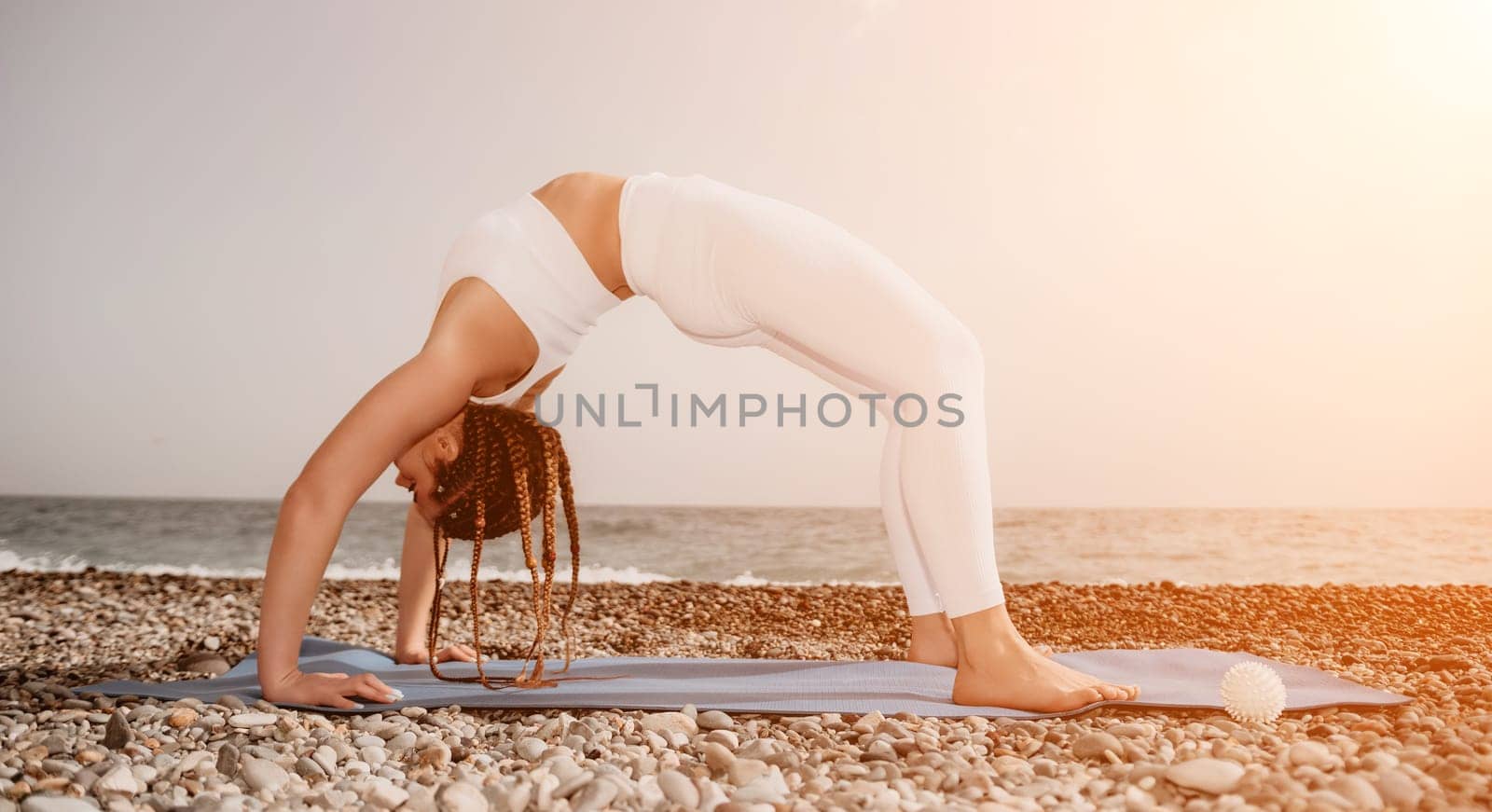 Woman yoga sea. Well looking middle aged woman with braids dreadlocks in white leggings and tops doing stretching pilates on yoga mat near sea. Female fitness yoga routine concept. Healthy lifestyle