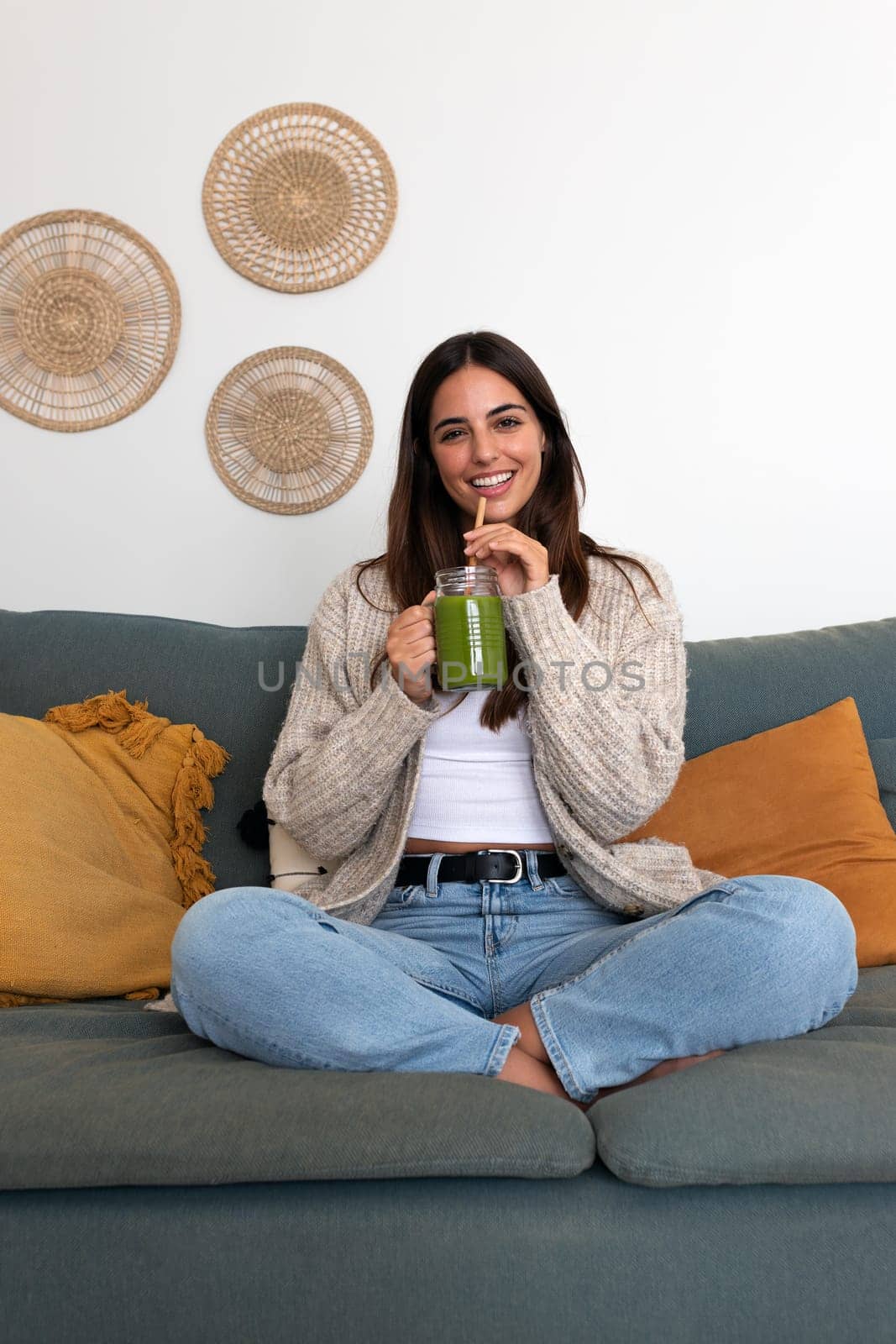 Vertical portrait of young beautiful woman relaxing at home sitting on the sofa drinking healthy green juice. Female enjoying smoothie looking at camera. Healthy lifestyle concept.