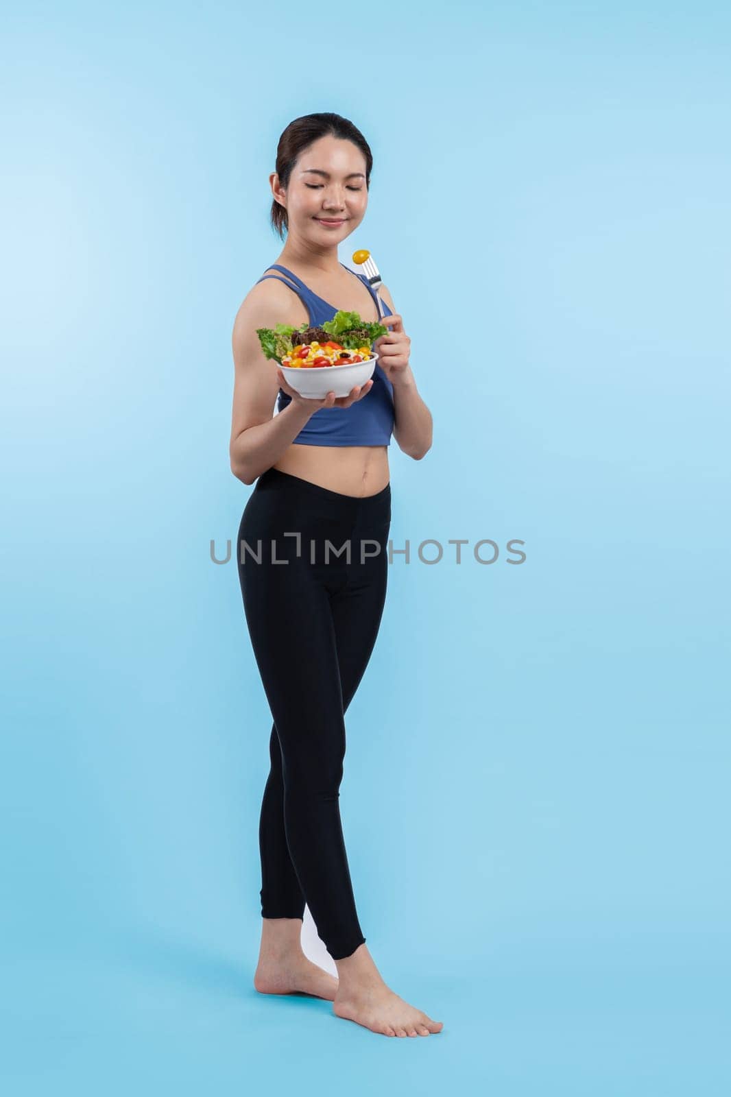 Young sporty Asian woman in sportswear holding salad bowl fill with vibrant of fruit and vegetable. Natural youthful and fit body lifestyle with balance nutrition on isolated background. Vigorous