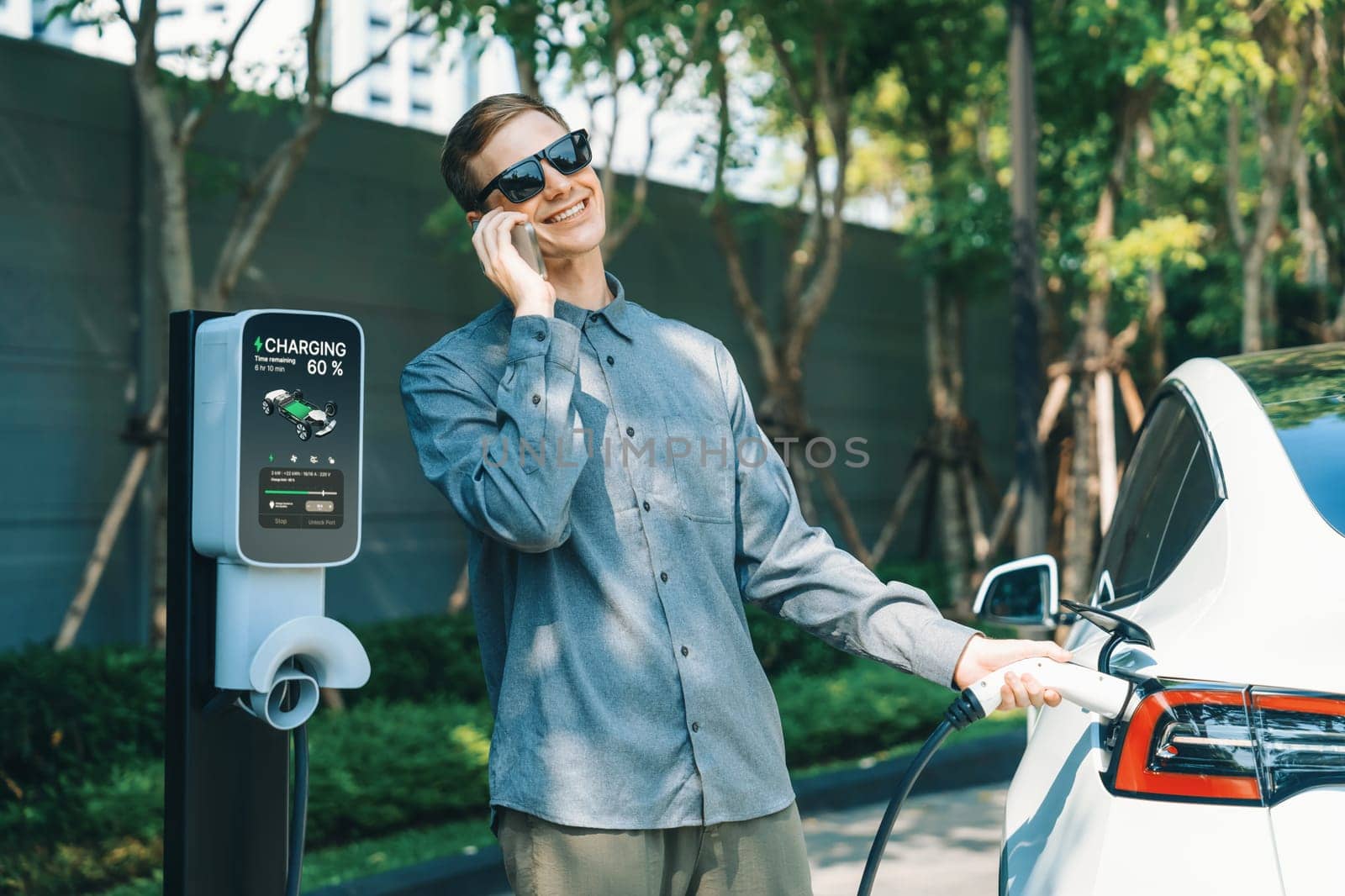 Young man travel with EV electric car charging in green sustainable city outdoor garden in summer shows urban sustainability lifestyle by green clean rechargeable energy of electric vehicle innards