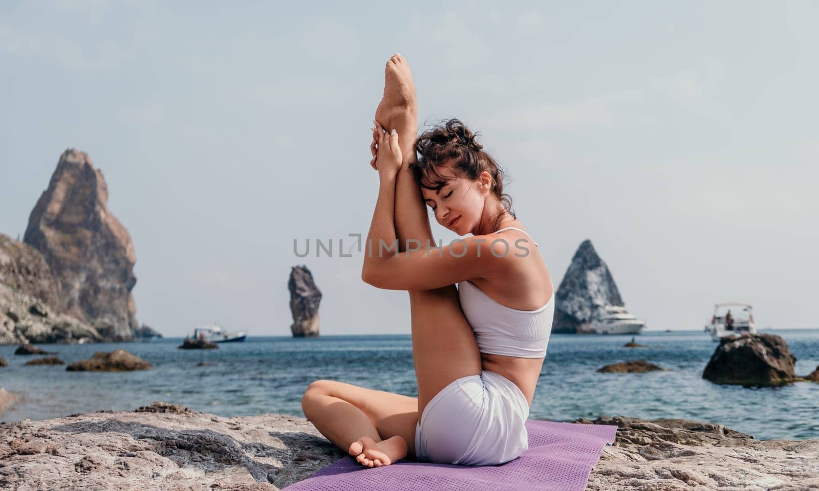 Fitness woman sea. A happy middle aged woman in white sportswear exercises morning outdoors on a beach with volcanic rocks by the sea. Female fitness pilates yoga routine concept. Healthy lifestyle. by panophotograph