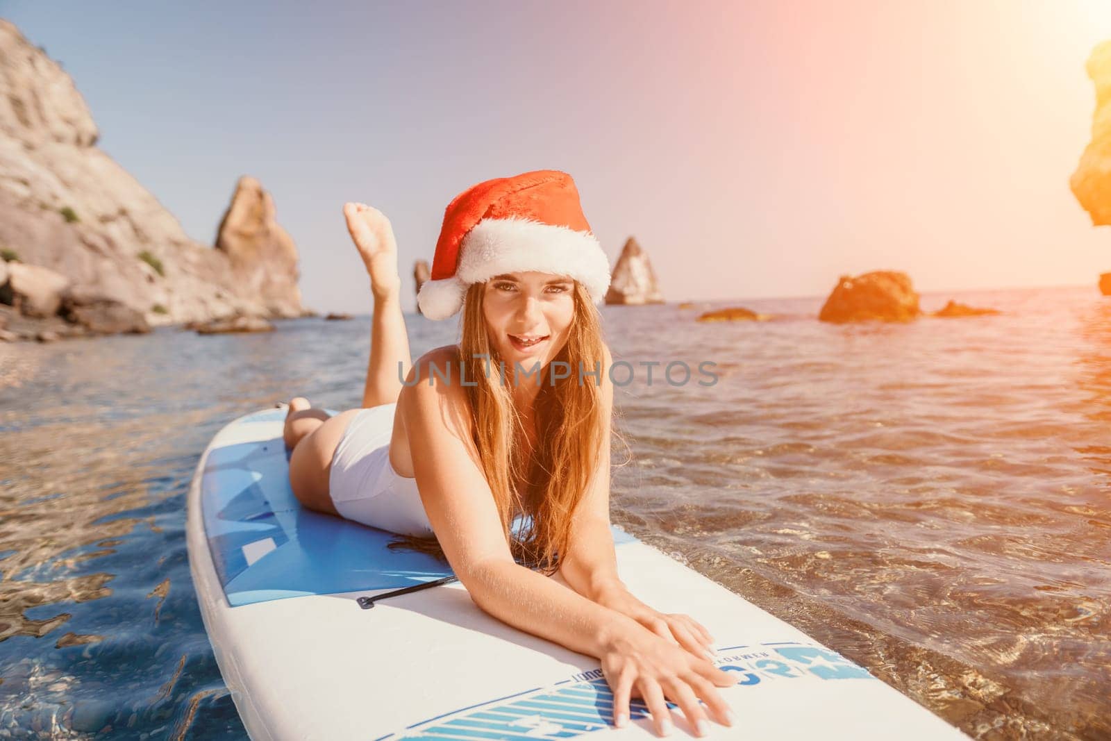 Woman sea sup. Close up portrait of happy young caucasian woman with long hair in Santa hat looking at camera and smiling. Cute woman portrait in a white bikini posing on sup board in the sea by panophotograph