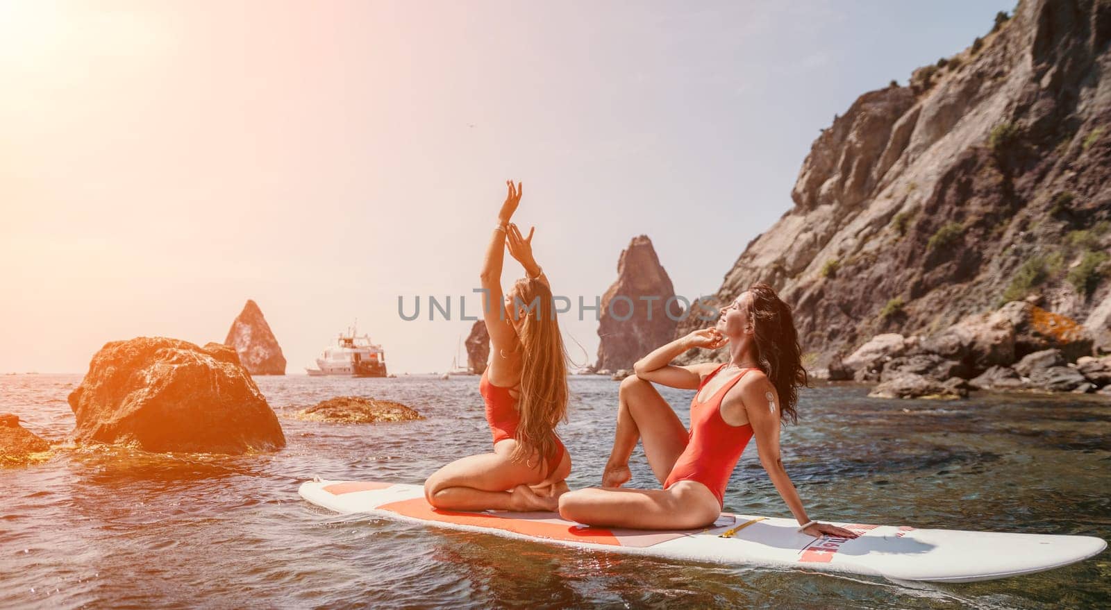 Woman sup yoga. Two happy sporty women practising yoga pilates on paddle sup surfboard. Female stretching doing workout on sea water. Modern individual female hipster outdoor summer sport activity. by panophotograph