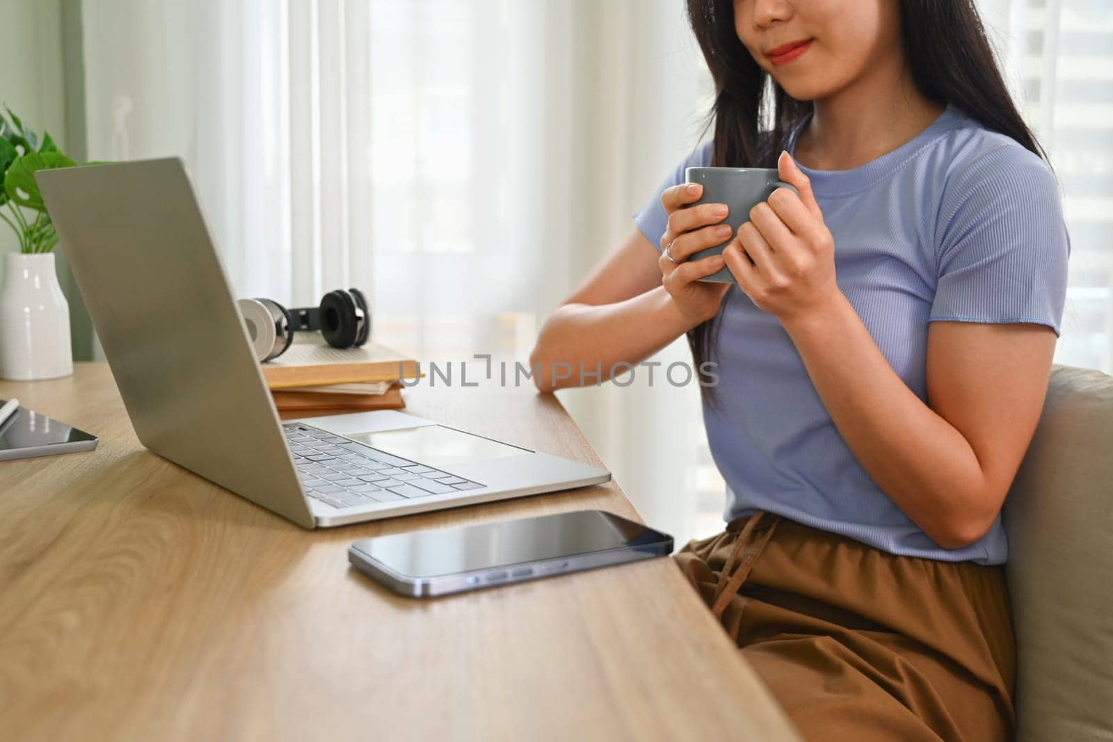Smiling young woman in casual clothes drinking tea and reading email or news on laptop computer.