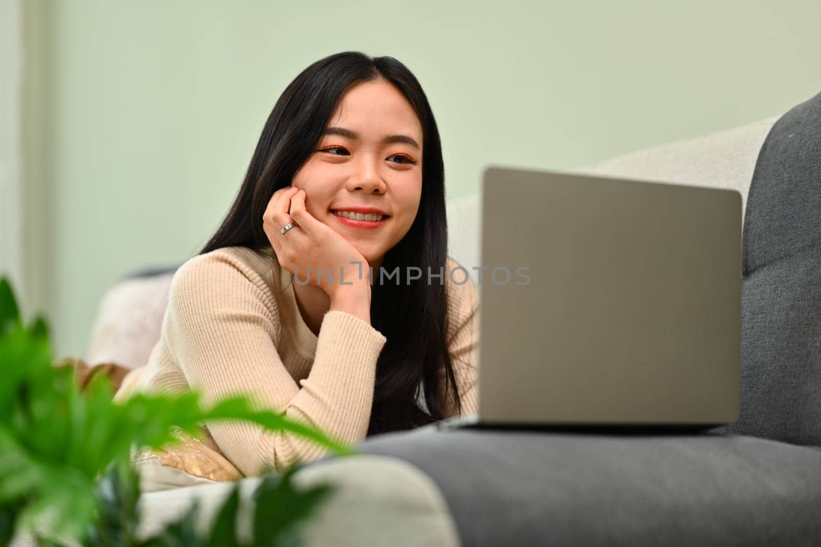 Happy young woman watching movies on laptop, lying on cozy sofa, enjoying leisure weekend time at home.