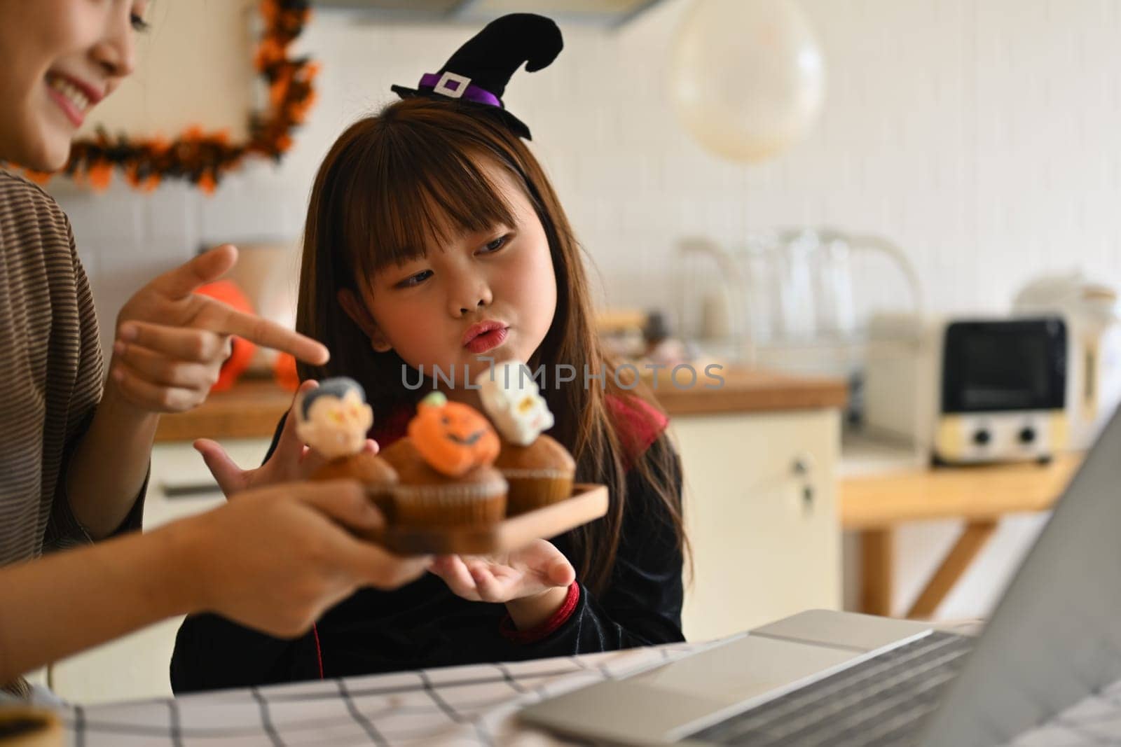 Playful little girl dressed as a witch and mother holding colorful Halloween cupcakes and video call on laptop in kitchen.