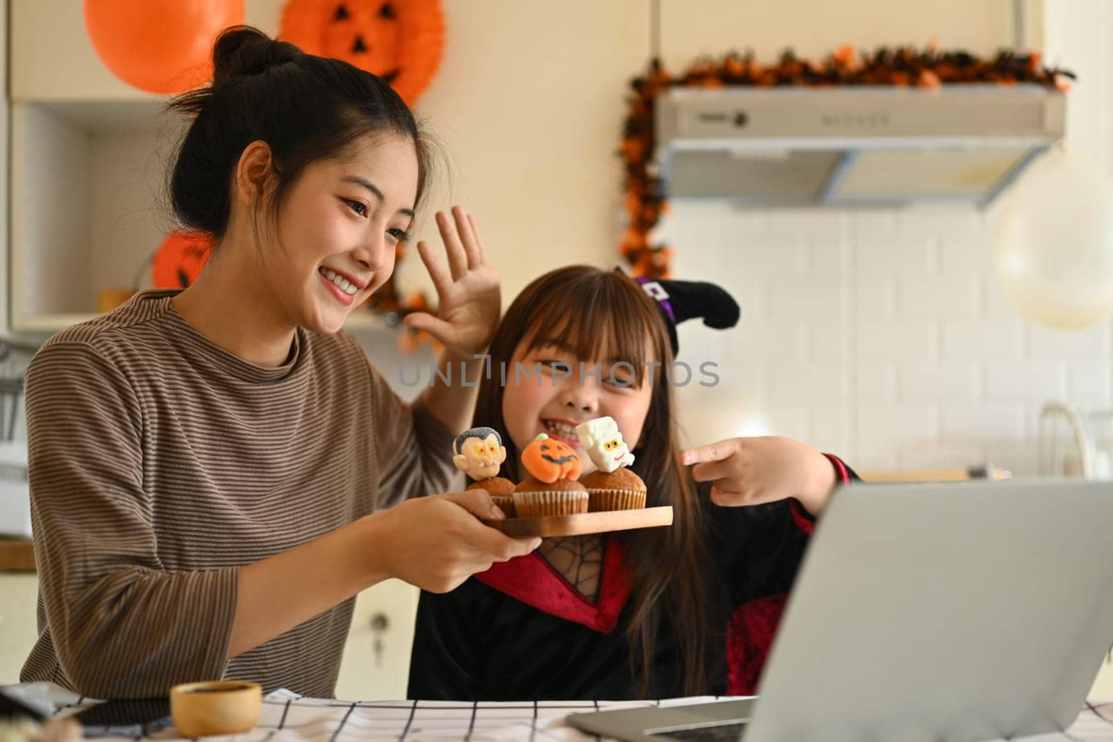 Playful little girl dressed as a witch and mother holding colorful Halloween cupcakes and video call on laptop in kitchen by prathanchorruangsak