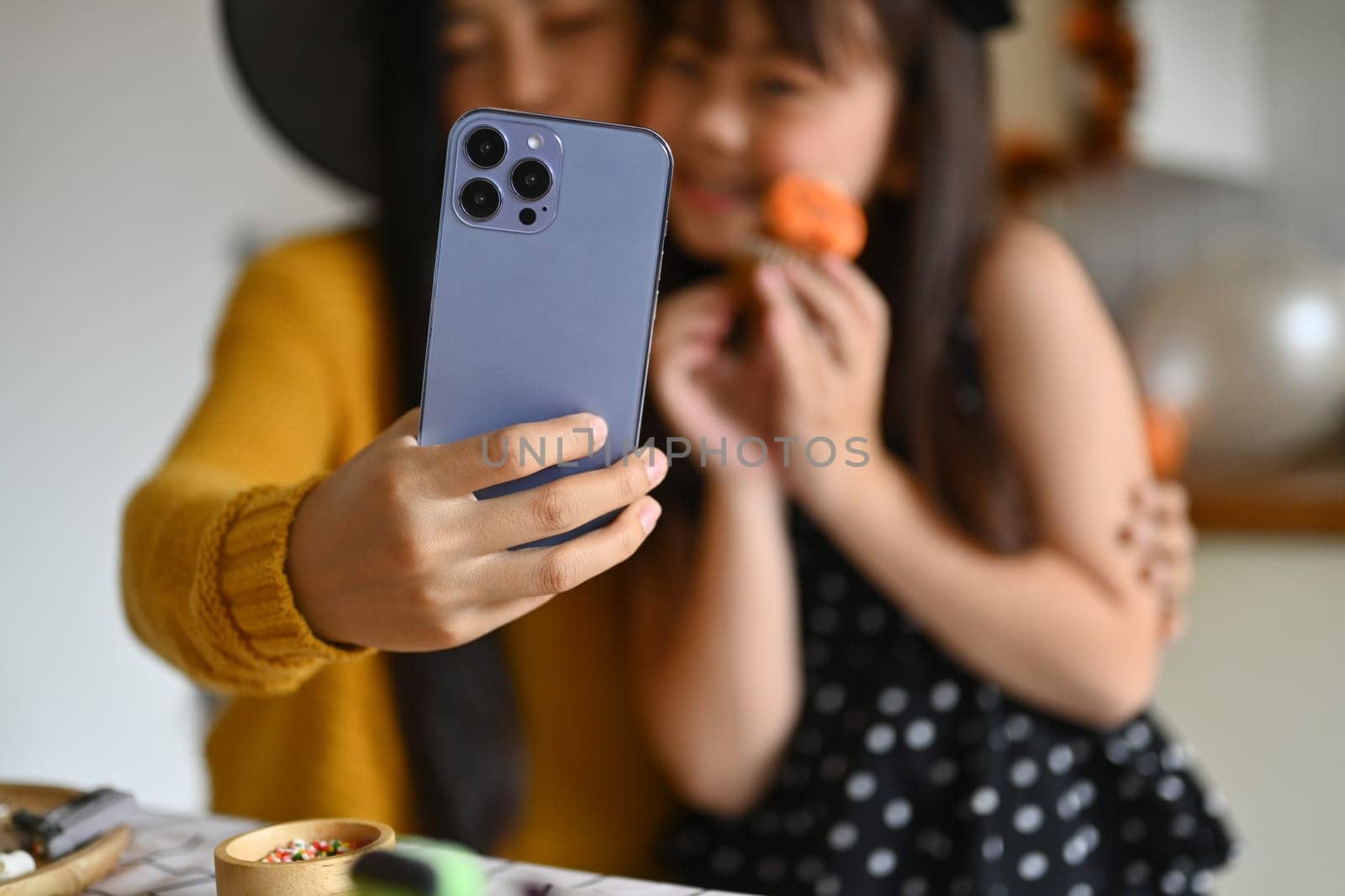 Smiling asian mother taking a selfie on smart phone and having fun with her daughter in Halloween decorated kitchen by prathanchorruangsak