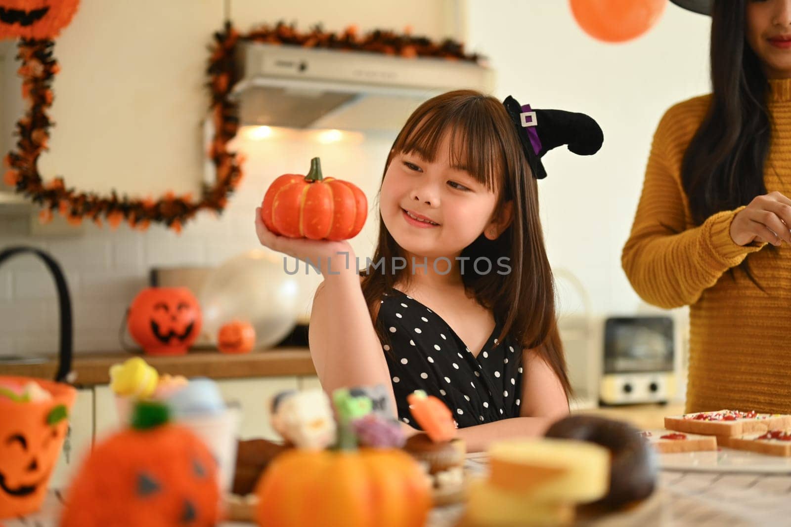 Adorable little Asian girl holding small pumpkin up by her face while preparing for holiday party with her mother in kitchen.