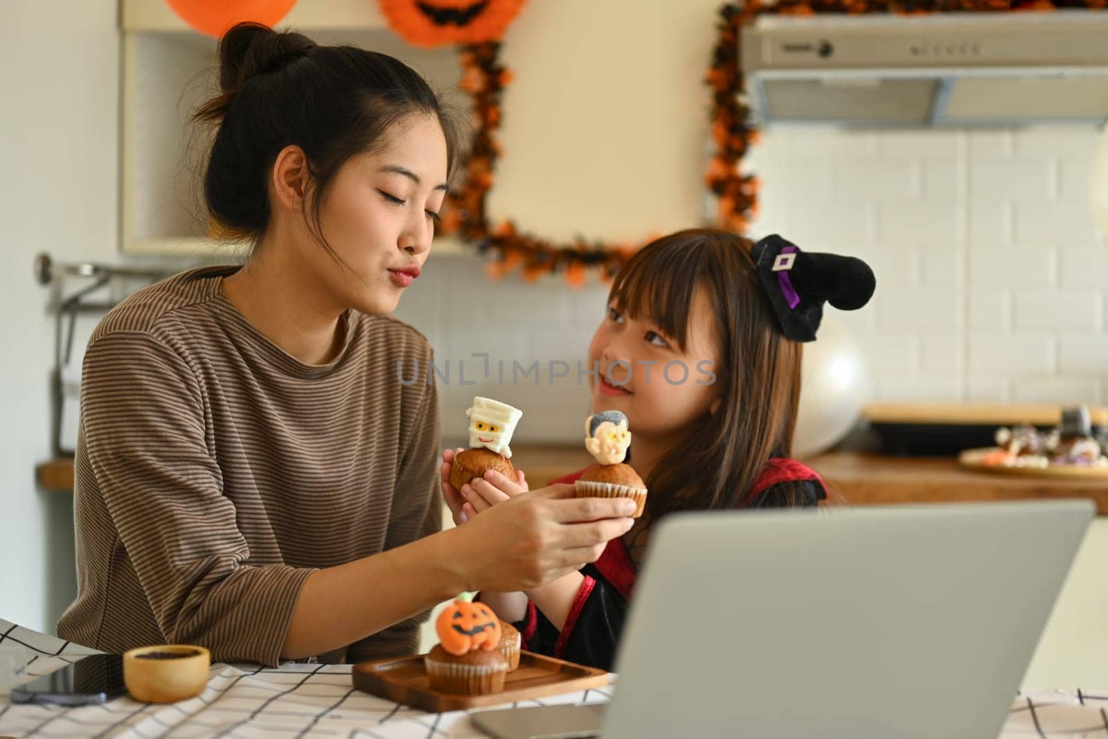 Playful little girl dressed as a witch and mother holding colorful Halloween cupcakes and video call on laptop in kitchen.
