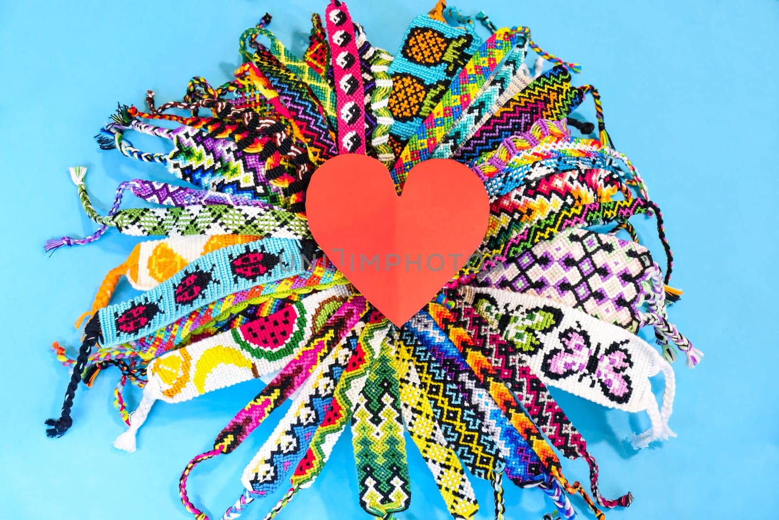 Red heart in the middle of circle of many multi-colored woven DIY friendship bracelets handmade of embroidery bright thread with knots on blue background.