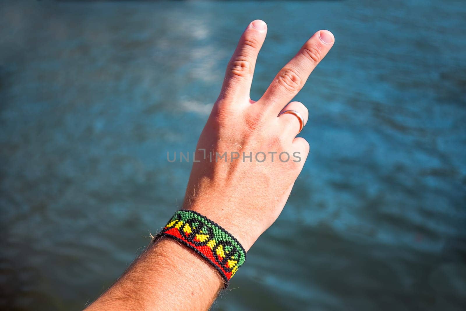 Selective focus of handmade DIY friendship bracelet with Rasta flag pattern with a peace symbol on male wrist on sea background.