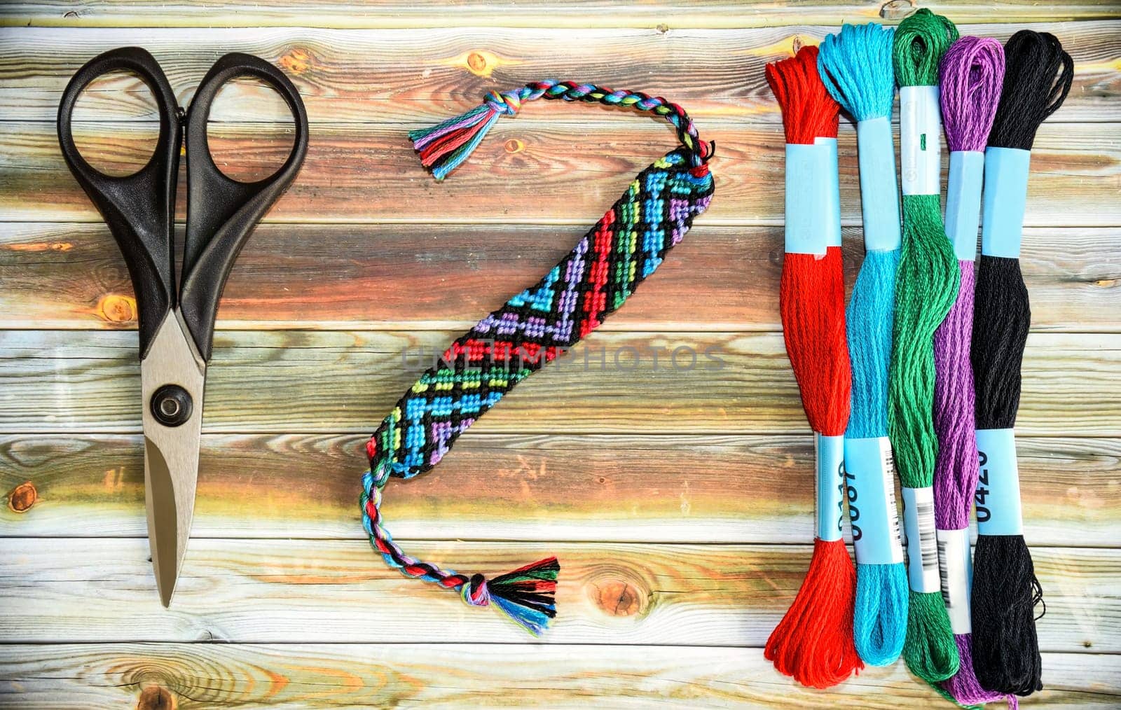 Woven DIY friendship bracelet with abstract pattern near skeins of embroidery floss, scissors on wooden background.