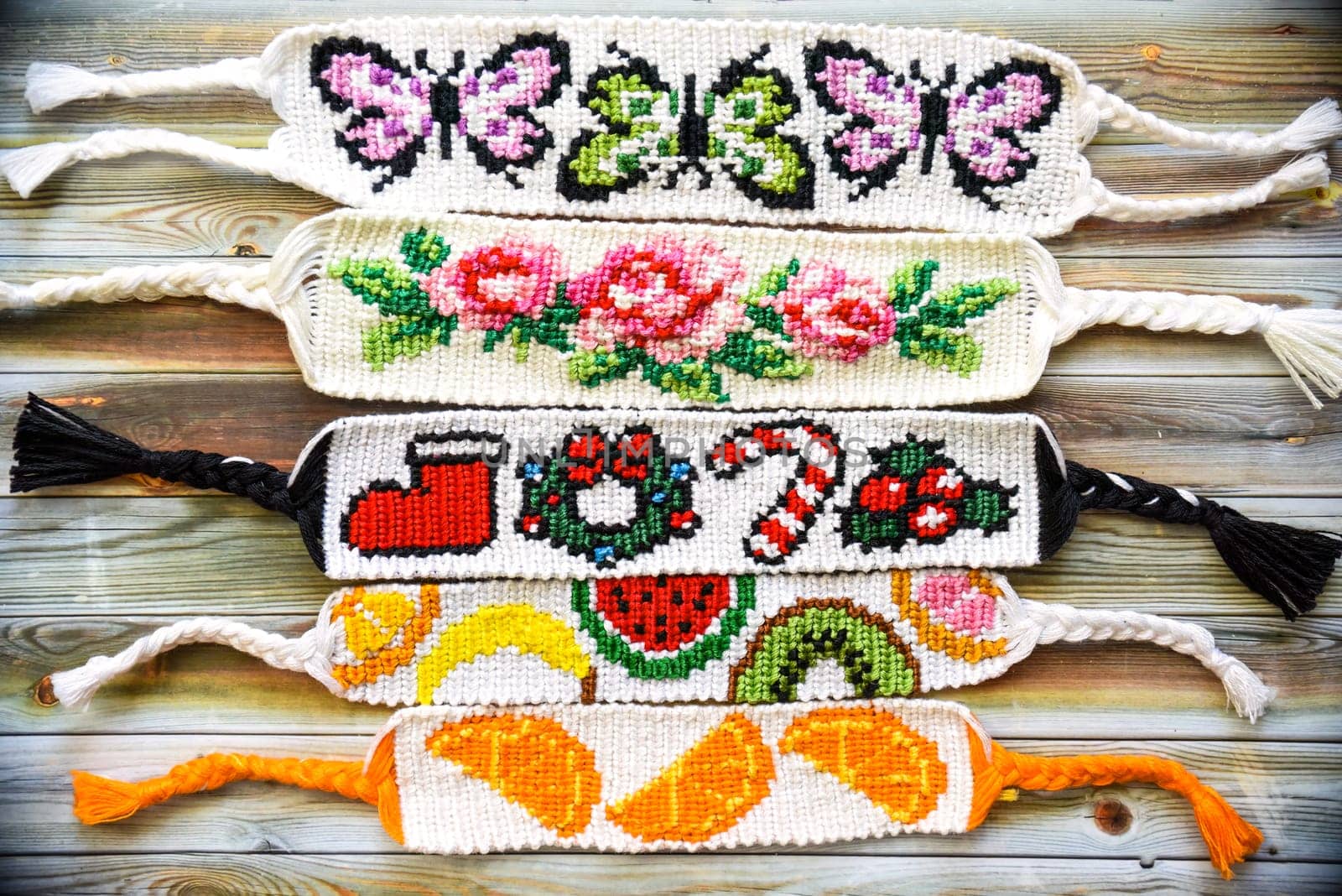 Woven colorful friendship bracelets with alpha patterns: butterflies, roses, christmas, oranges and fruits. Handmade of thread on wooden background. DIY gift idea