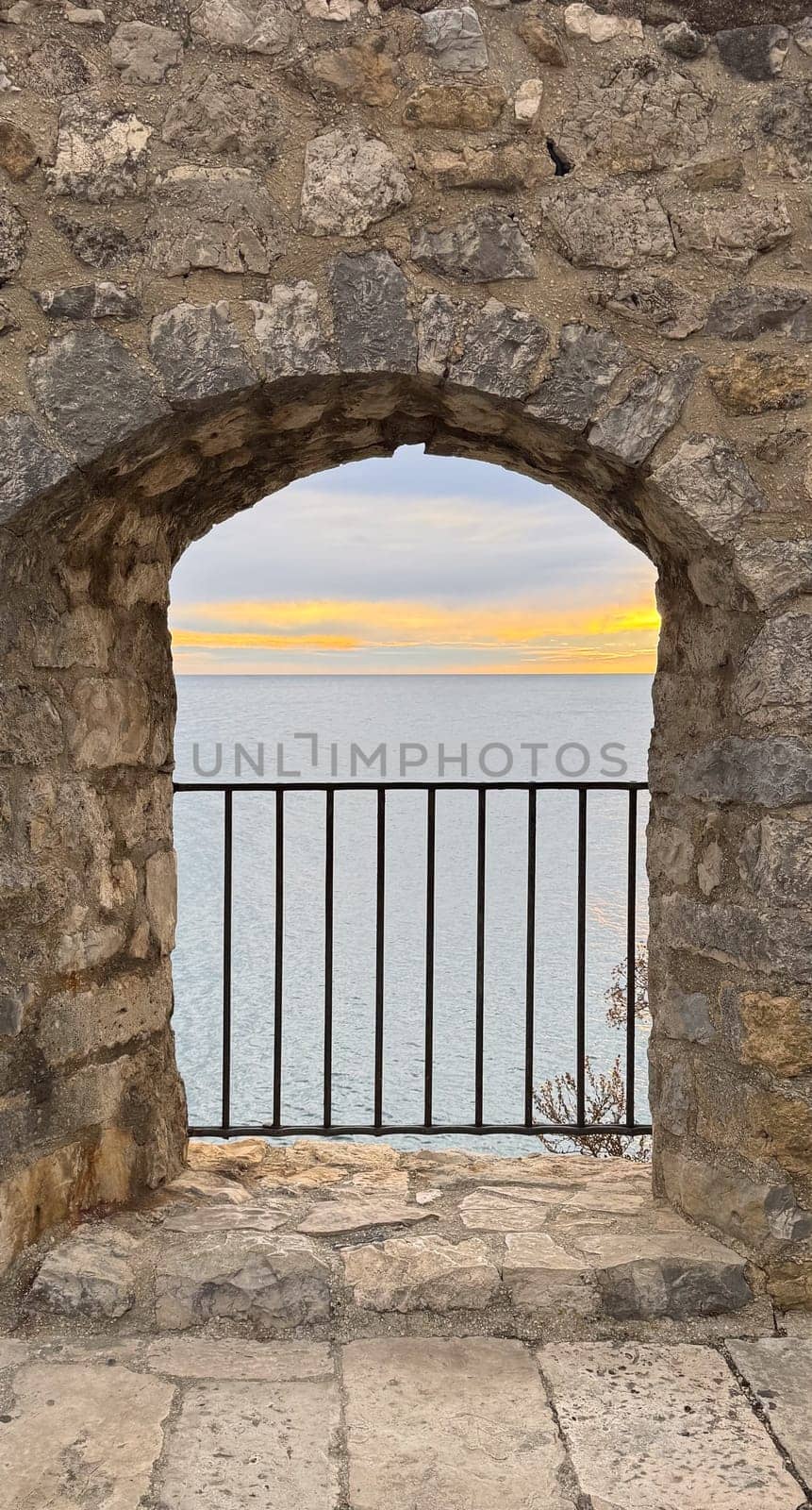 A rustic stone archway frames a view of calm blue waters and a golden sunset, with a black iron railing in the foreground. High quality photo